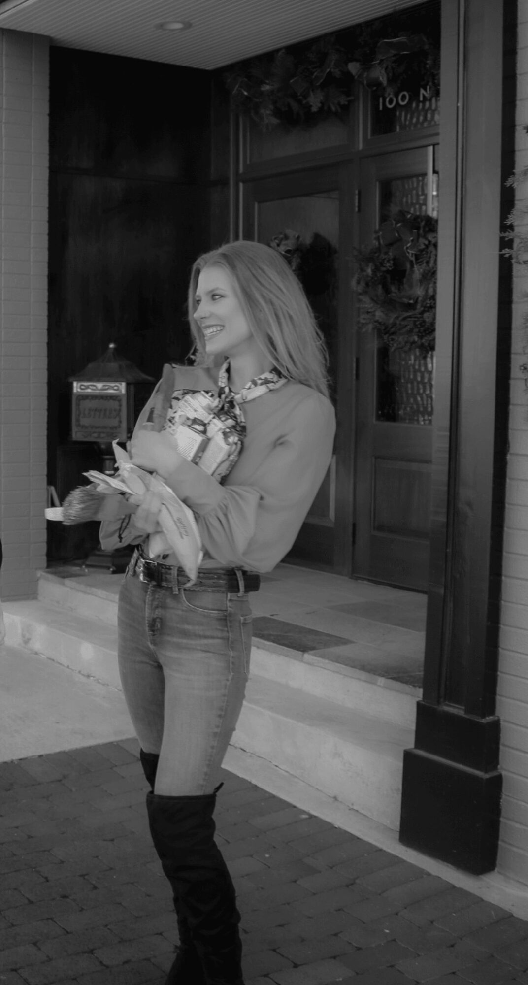 Woman holding a bouquet of flowers standing outside a building entrance, smiling, with a door and window behind her.