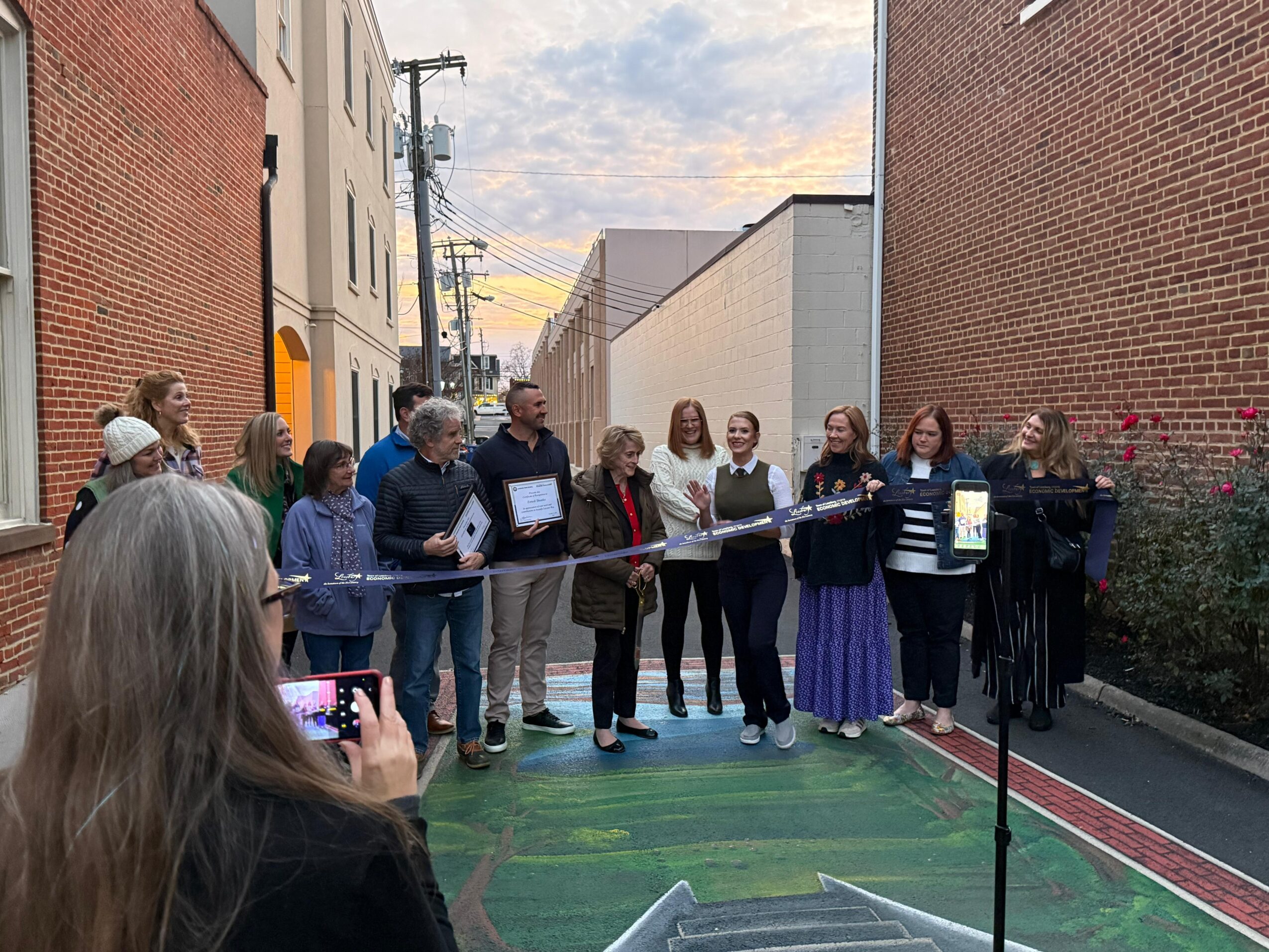 Group of people standing on a colorful outdoor court, some holding papers, with a person taking a photo.