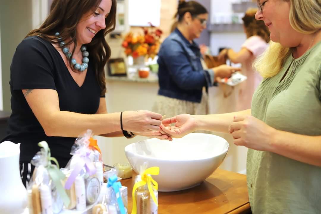 Two women exchanging a ring in a brightly lit room with a large white bowl on the table, others in the background.