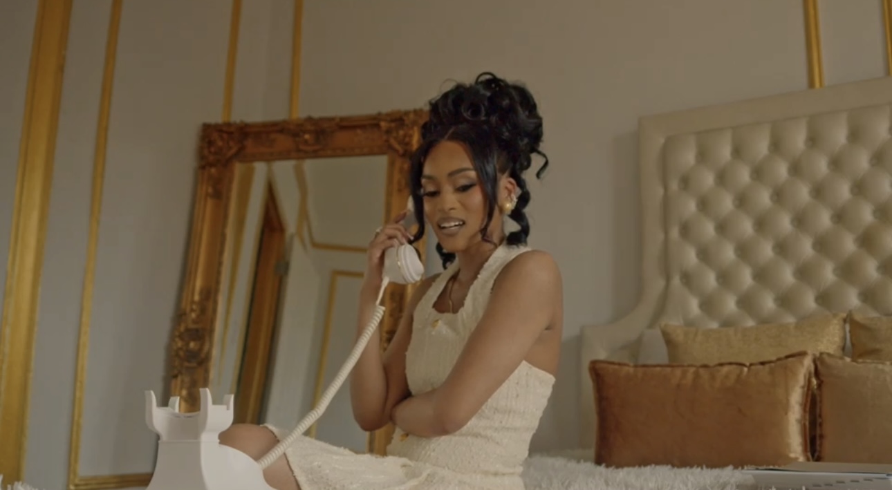 Woman in white dress talking on a vintage telephone in a decorated bedroom with a large mirror and bed.
