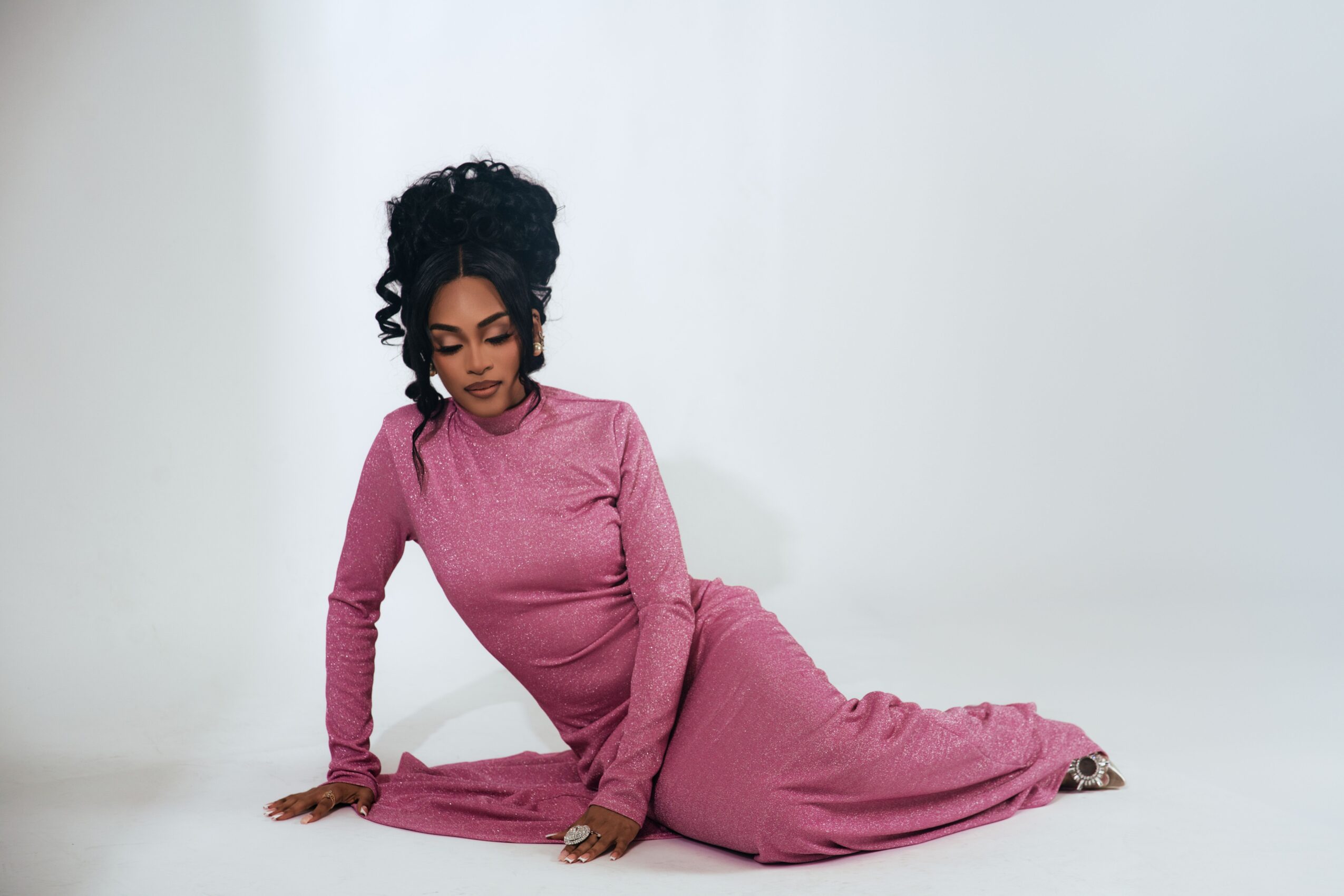 Woman in pink dress sitting on the floor against a plain background, looking down, with curly hair styled up.