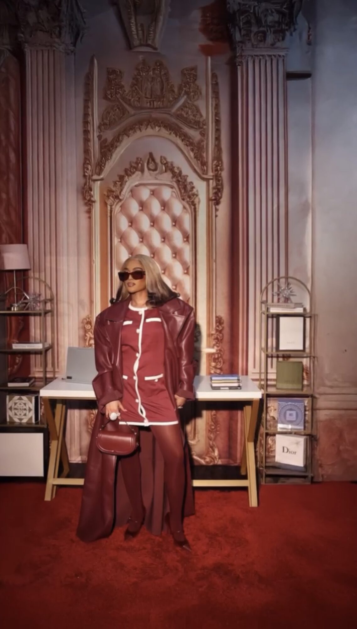Person with blonde hair and sunglasses standing in front of ornate pink and gold backdrop, wearing a red leather jacket and pants.