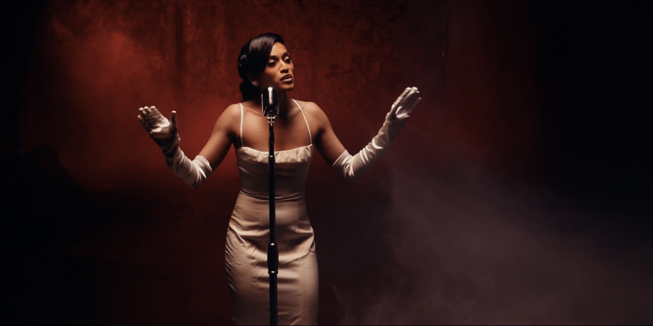 A woman in a beige dress and gloves sings into a microphone with a red and black background.