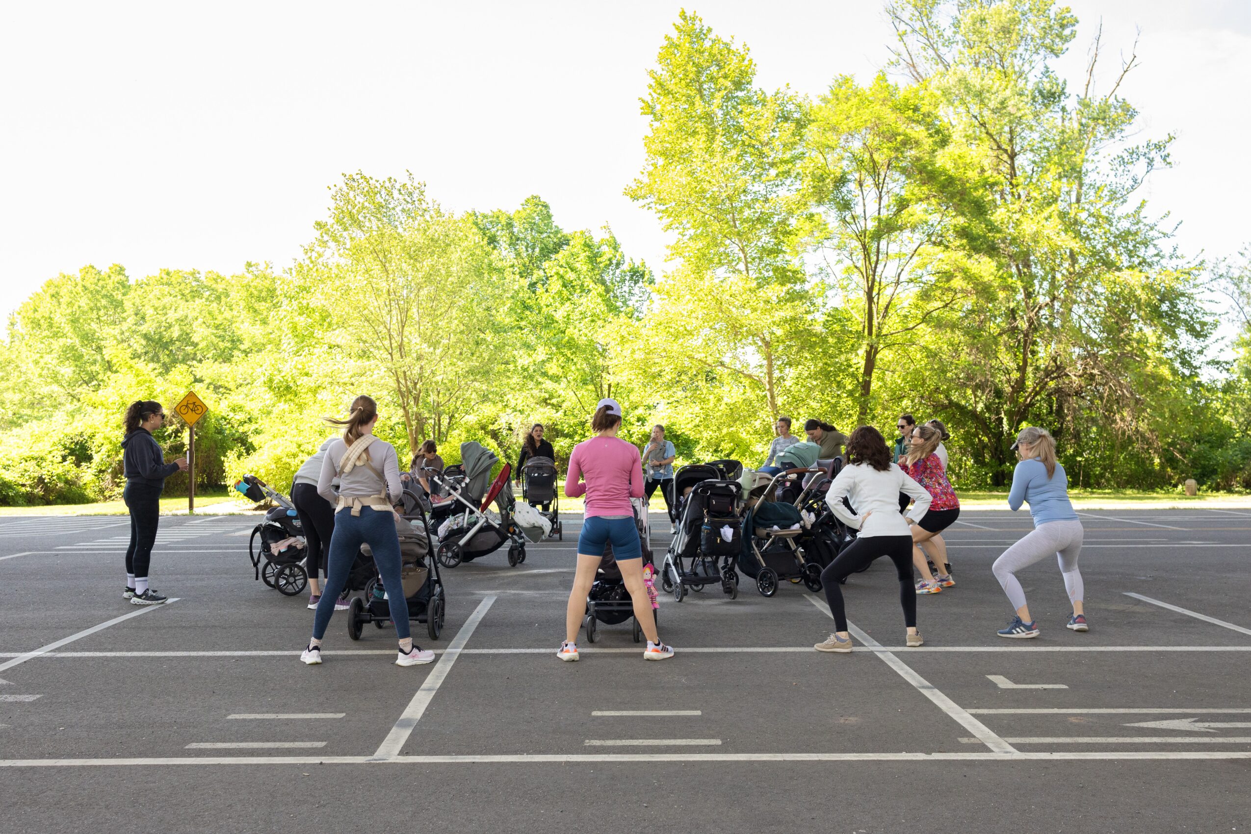 Group of people in a parking lot with trees in the background, some standing and others sitting or leaning near motorcycles.
