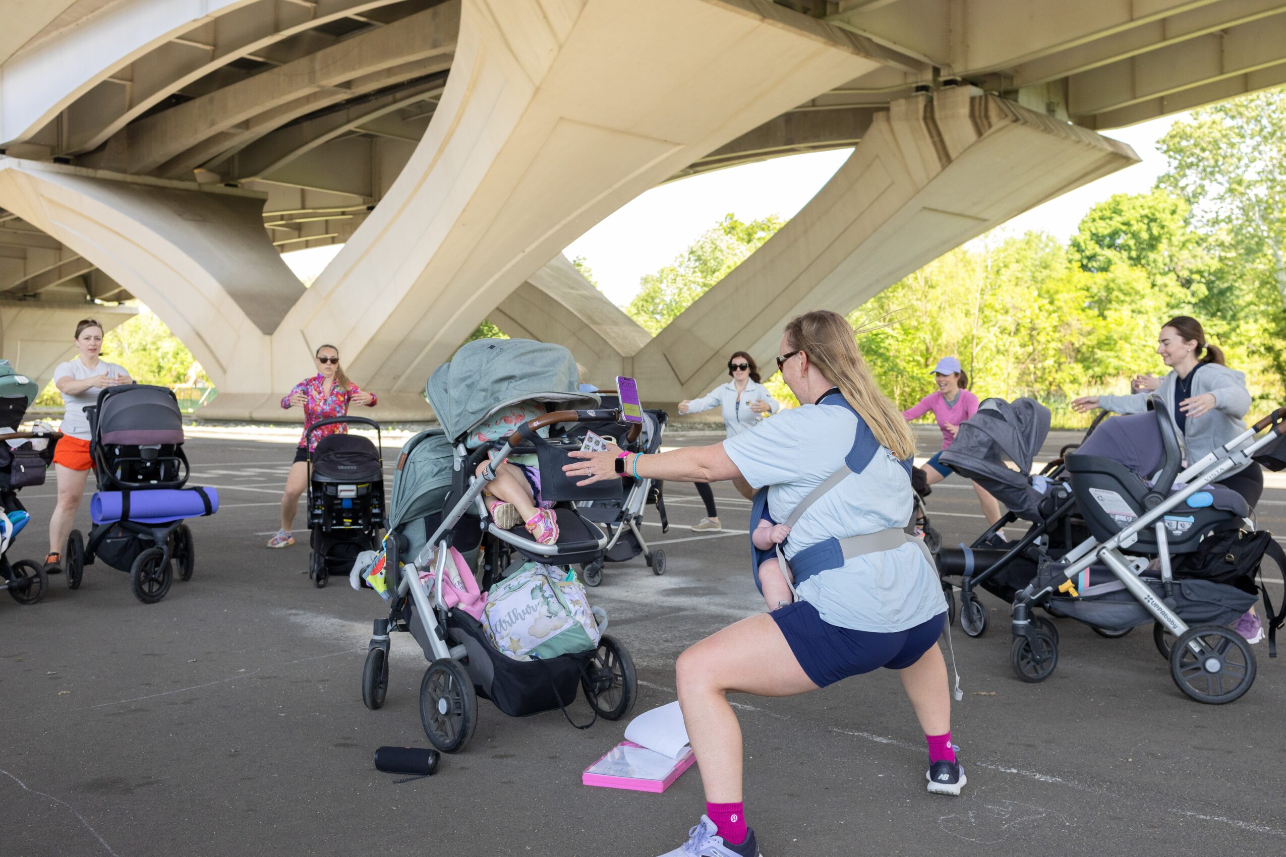 Group of people with strollers under a bridge, some interacting, outdoors with trees in the background.