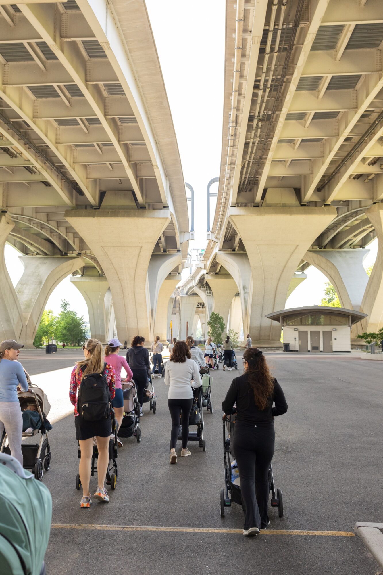 People walking under large elevated highways with supporting columns and a small building nearby, clear sky above.