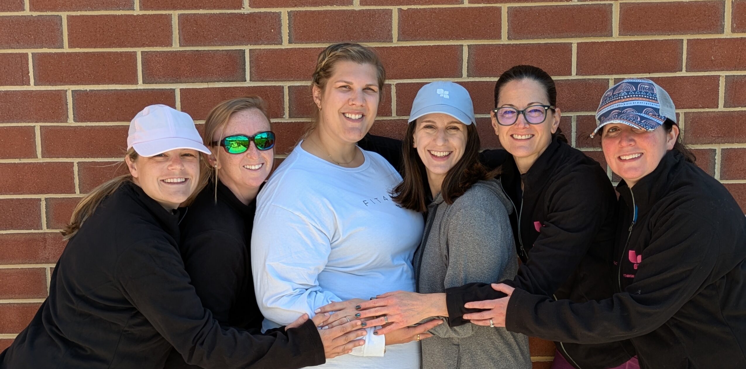 Six women standing against a brick wall, smiling and holding a piece of paper, wearing casual clothing and hats.