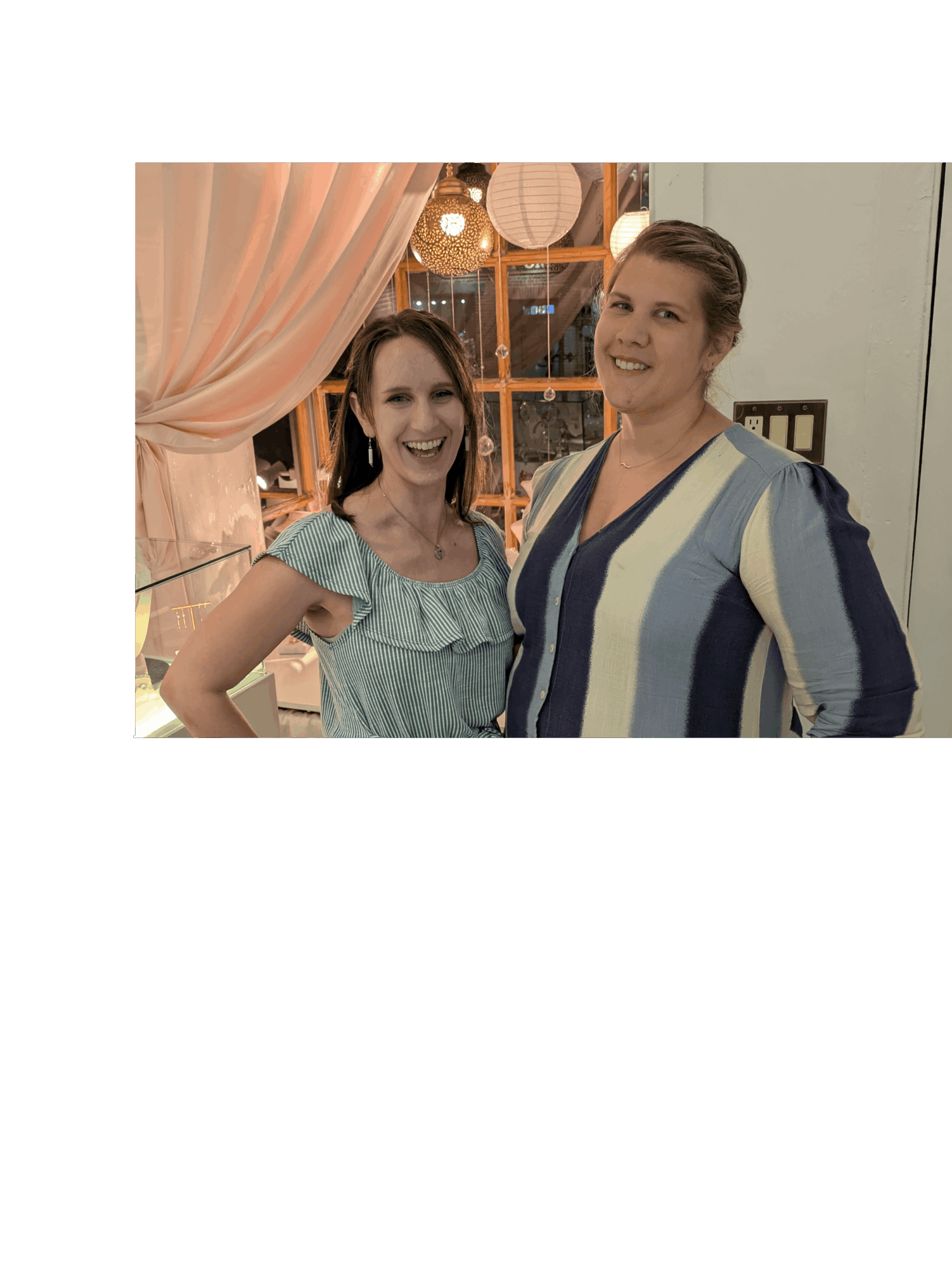 Two women smiling indoors, one wearing a light blue top, the other in a striped shirt, with pink curtains and warm lighting.