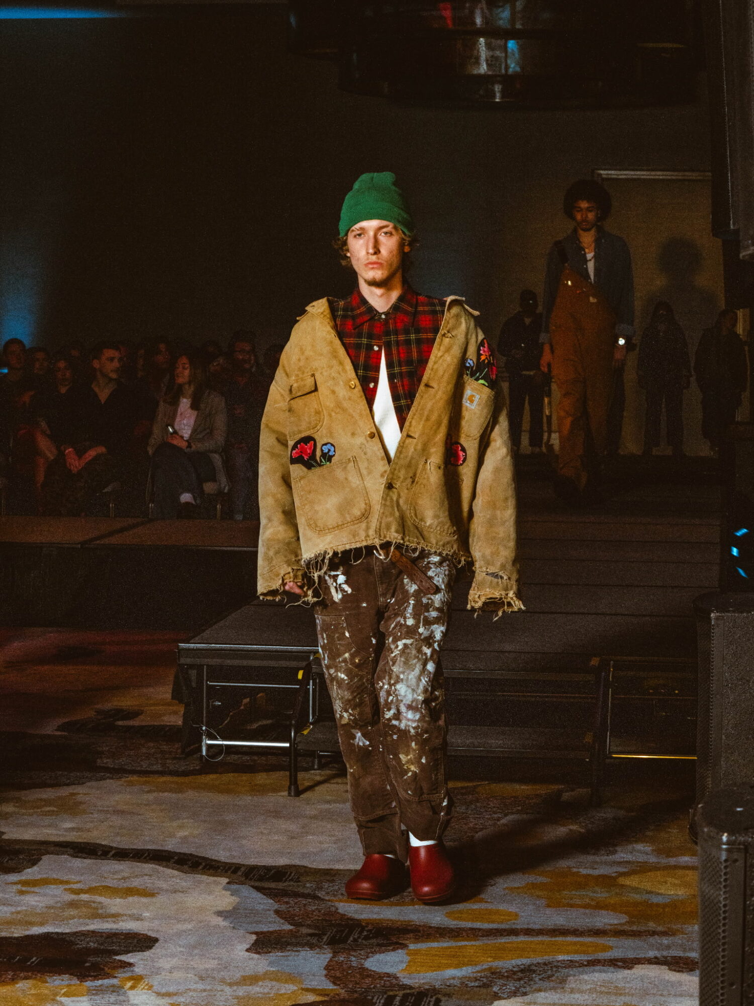 Person walking on runway wearing a green beanie, oversized beige jacket, patterned pants, and red shoes, with audience in background.