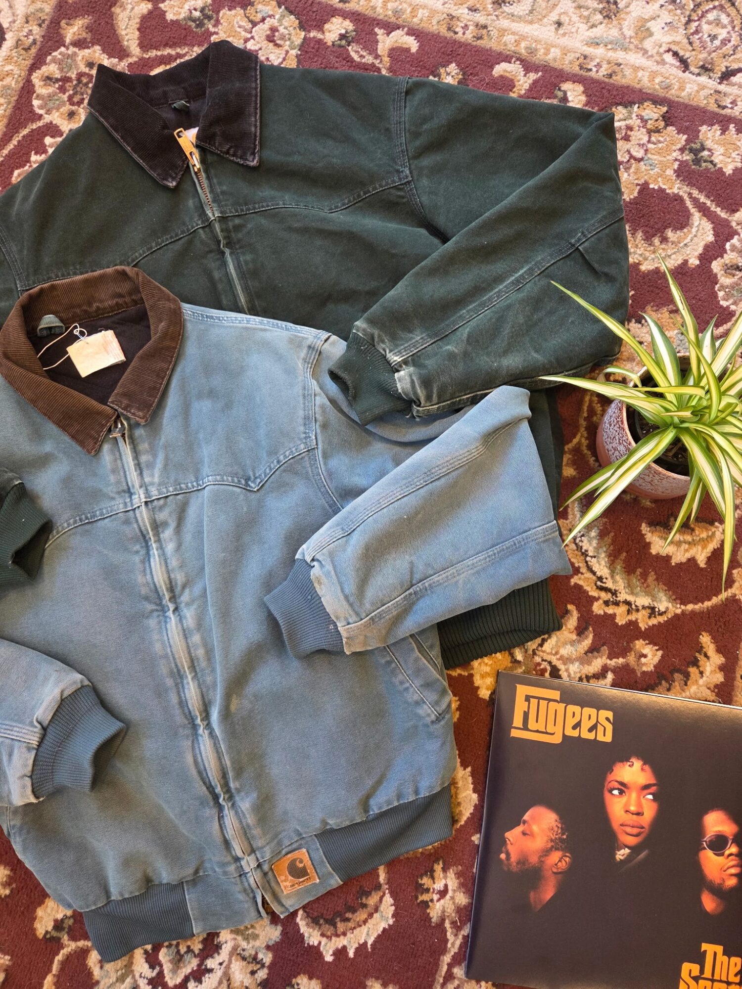 Two denim jackets, one dark and one light, are laid on a patterned rug with a potted plant nearby and a vinyl record cover.