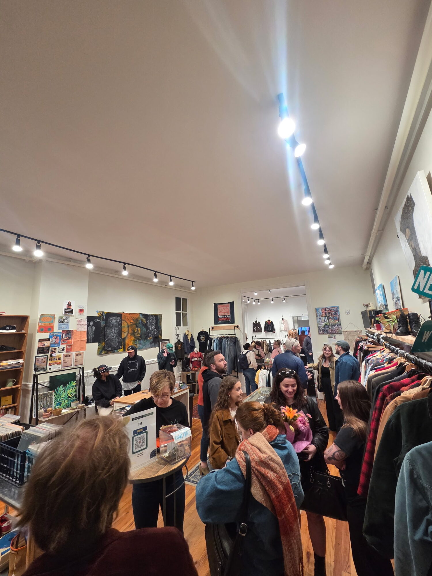 Crowded store with people shopping and browsing clothing racks, shelves, and displays, with bright lighting and artwork on walls.