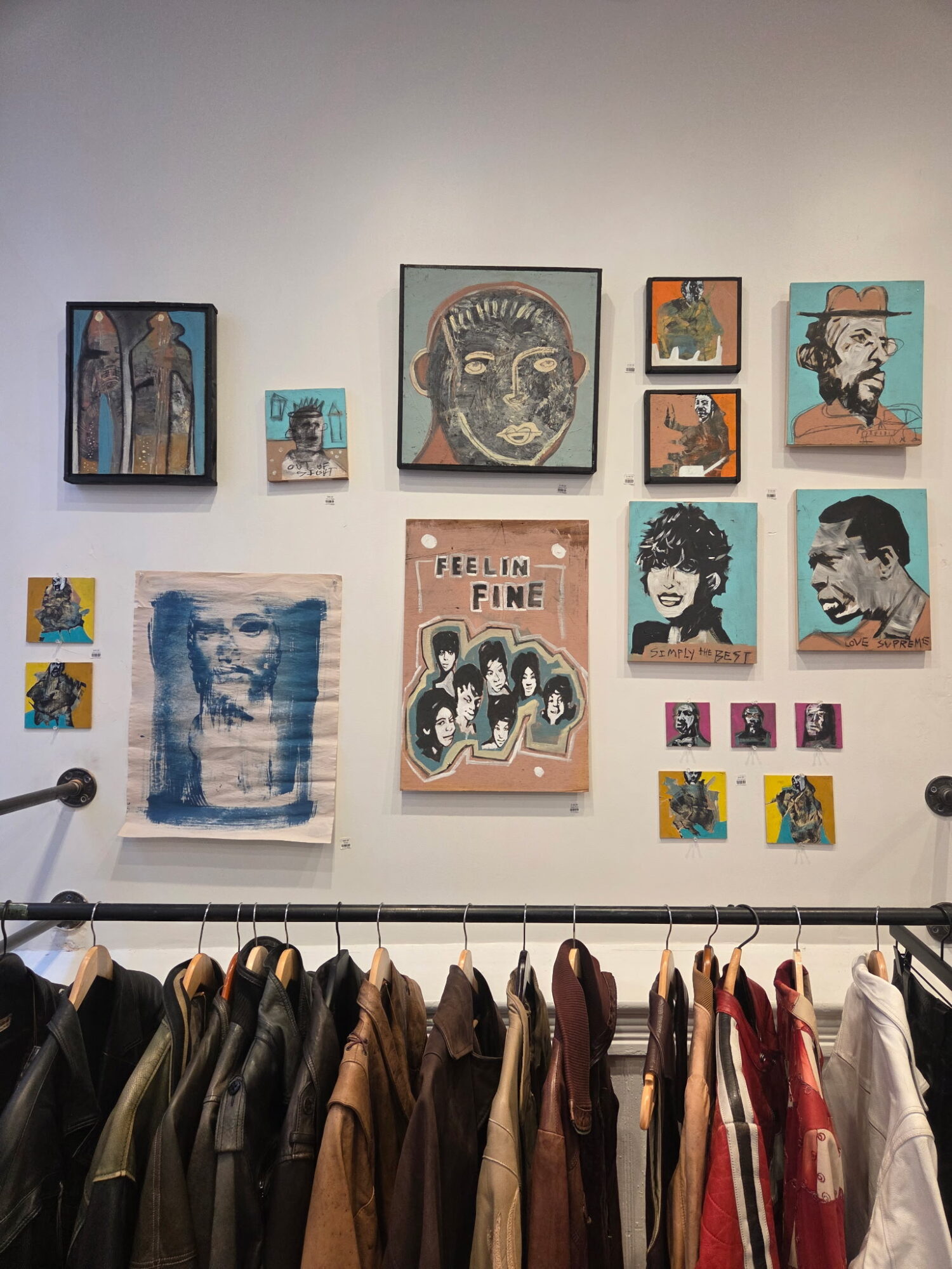 Gallery wall with various framed artworks, including portraits, abstract pieces, and a poster, above a clothing rack with jackets and shirts.