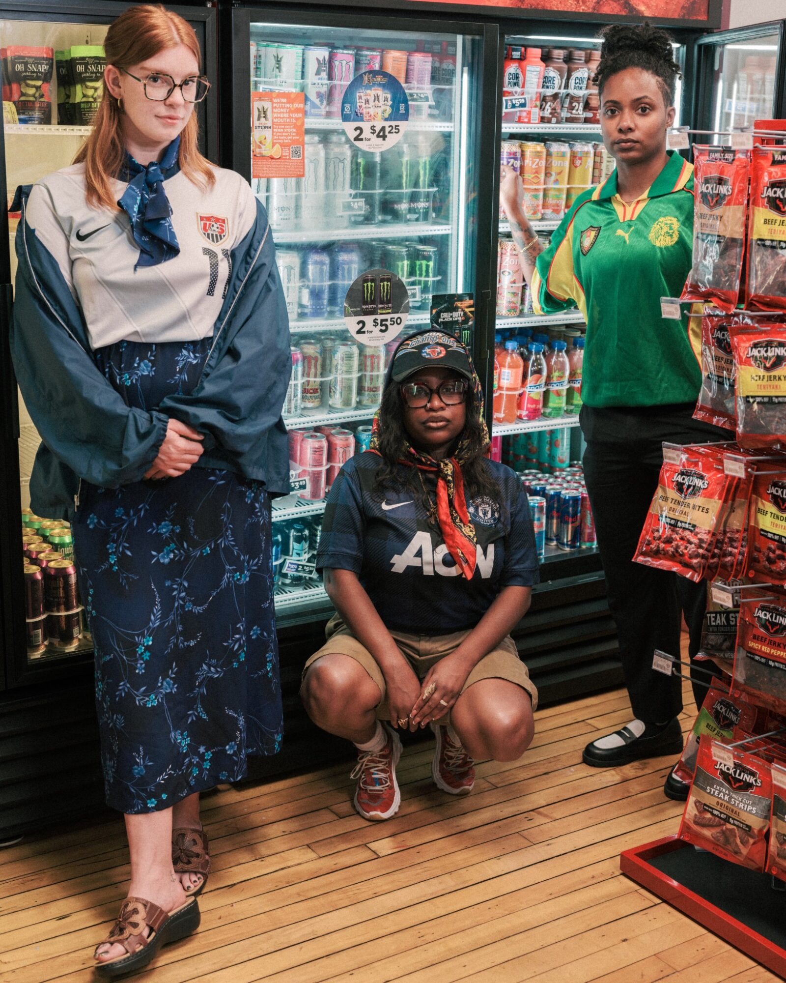 Three women in a store aisle, one sitting and two standing, with shelves of snacks and drinks behind them.