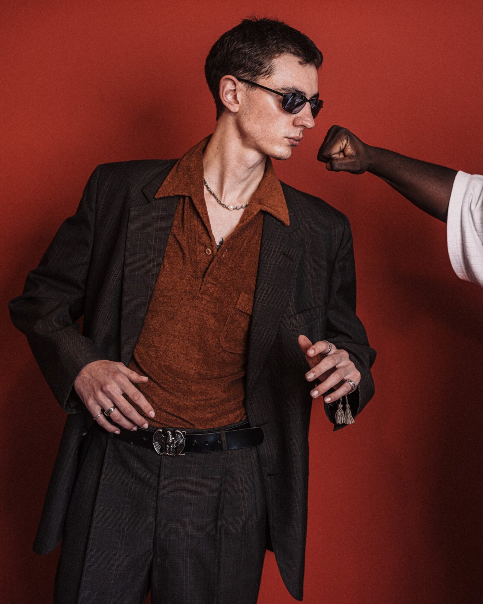 Person in sunglasses and suit with a brown shirt, standing against a red background, with a gloved hand reaching towards them.
