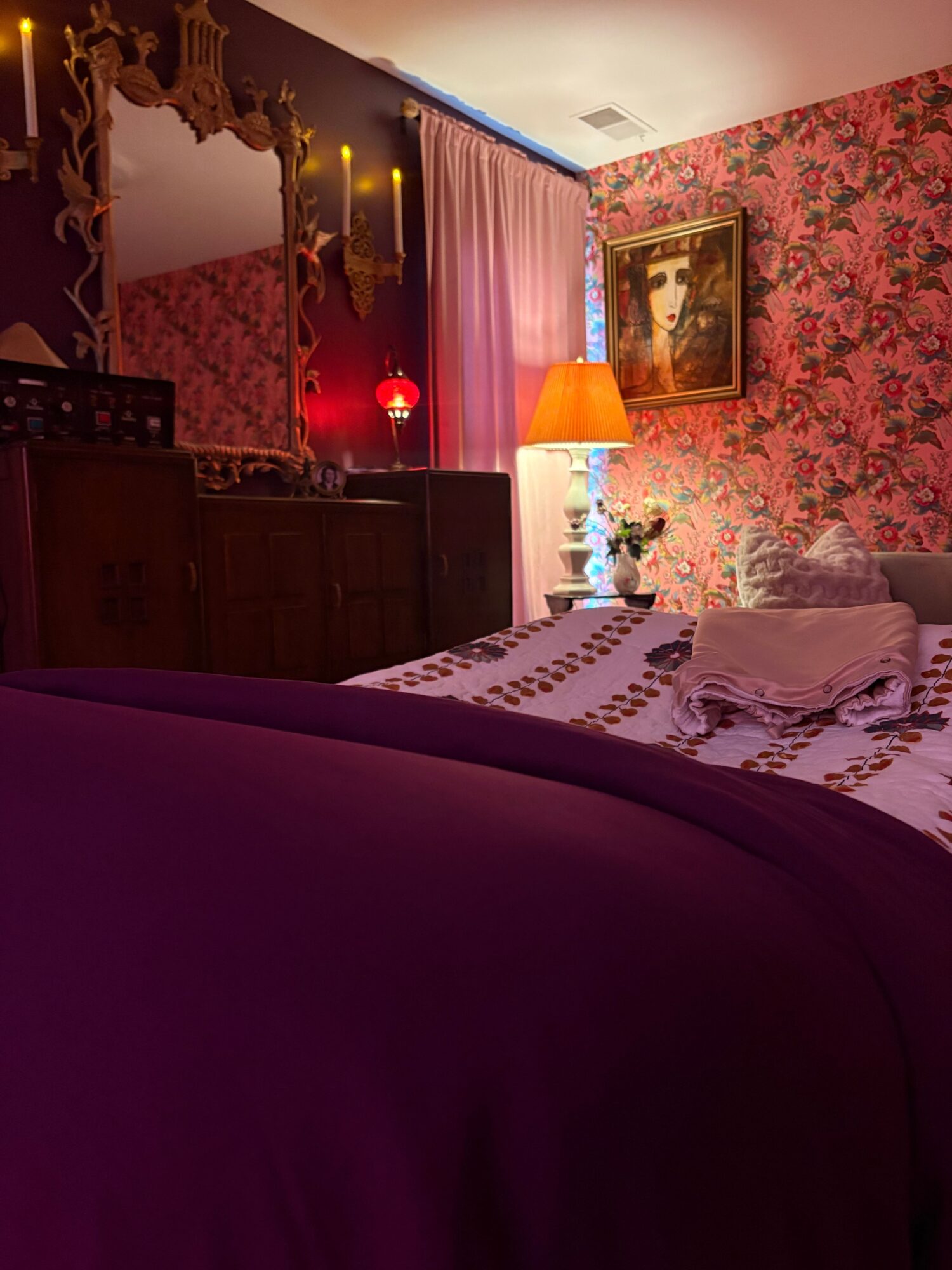 A bedroom with floral wallpaper, a bed with patterned bedding, a wooden dresser, and a window with curtains.