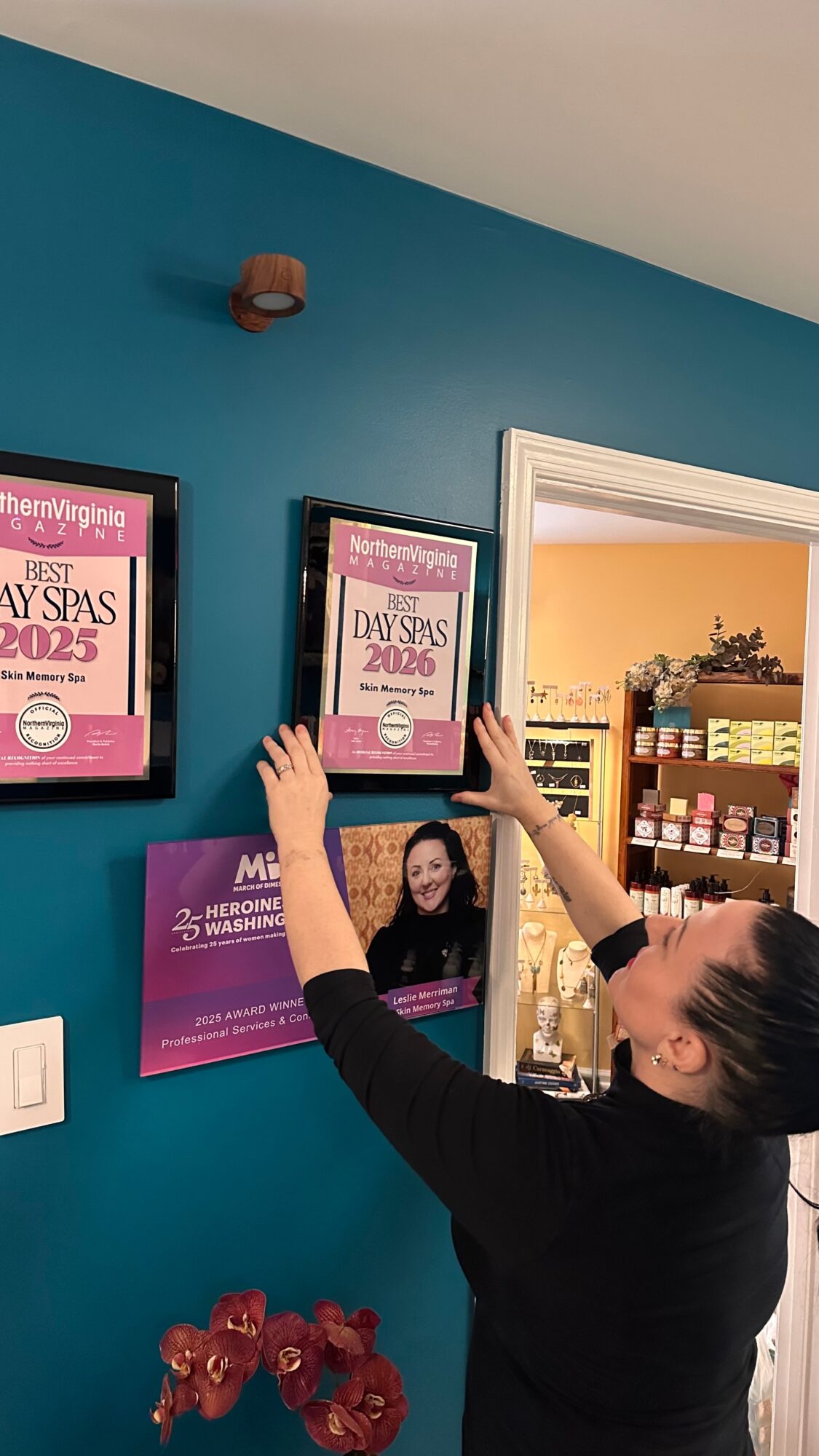 Woman hanging framed awards on a teal wall in a room with shelves and products, with a white ceiling above.