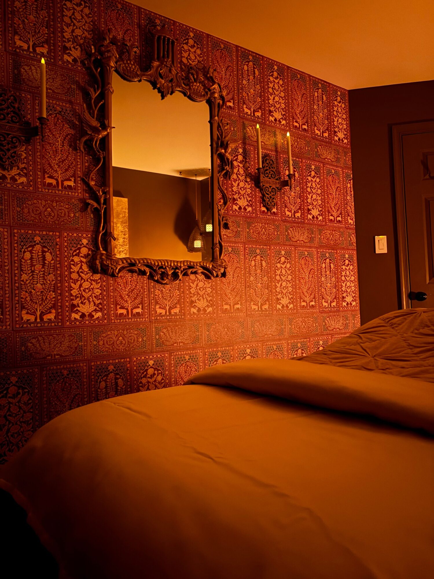 Decorative wall with a large ornate mirror and patterned wallpaper, partially visible bed on the right side.