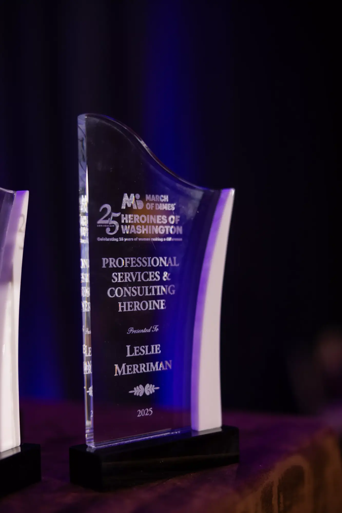 Glass award with text, on a black base, against a dark background, illuminated by purple light.