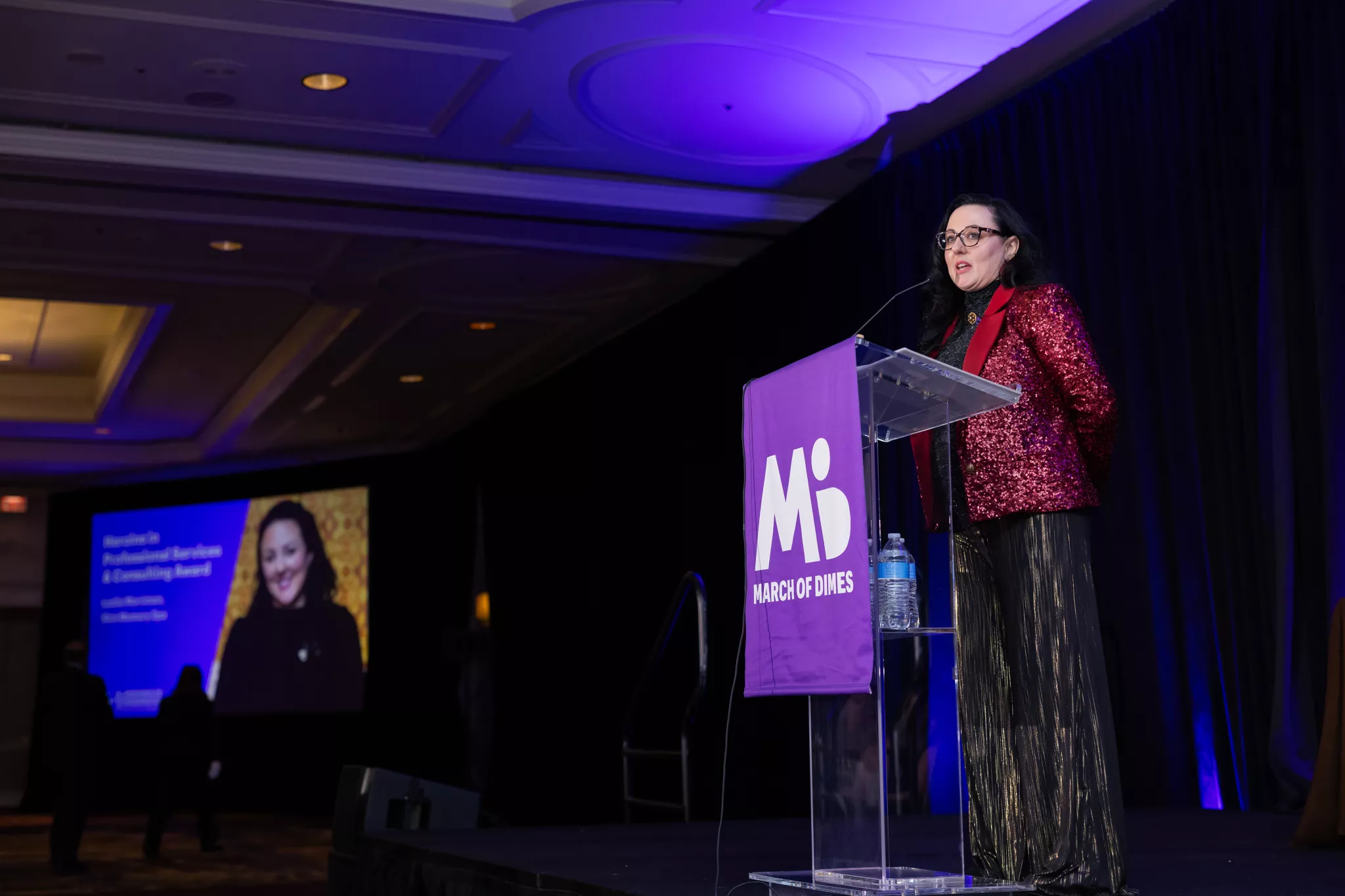 Woman speaking at a podium during a conference, with a large screen displaying her image in the background.