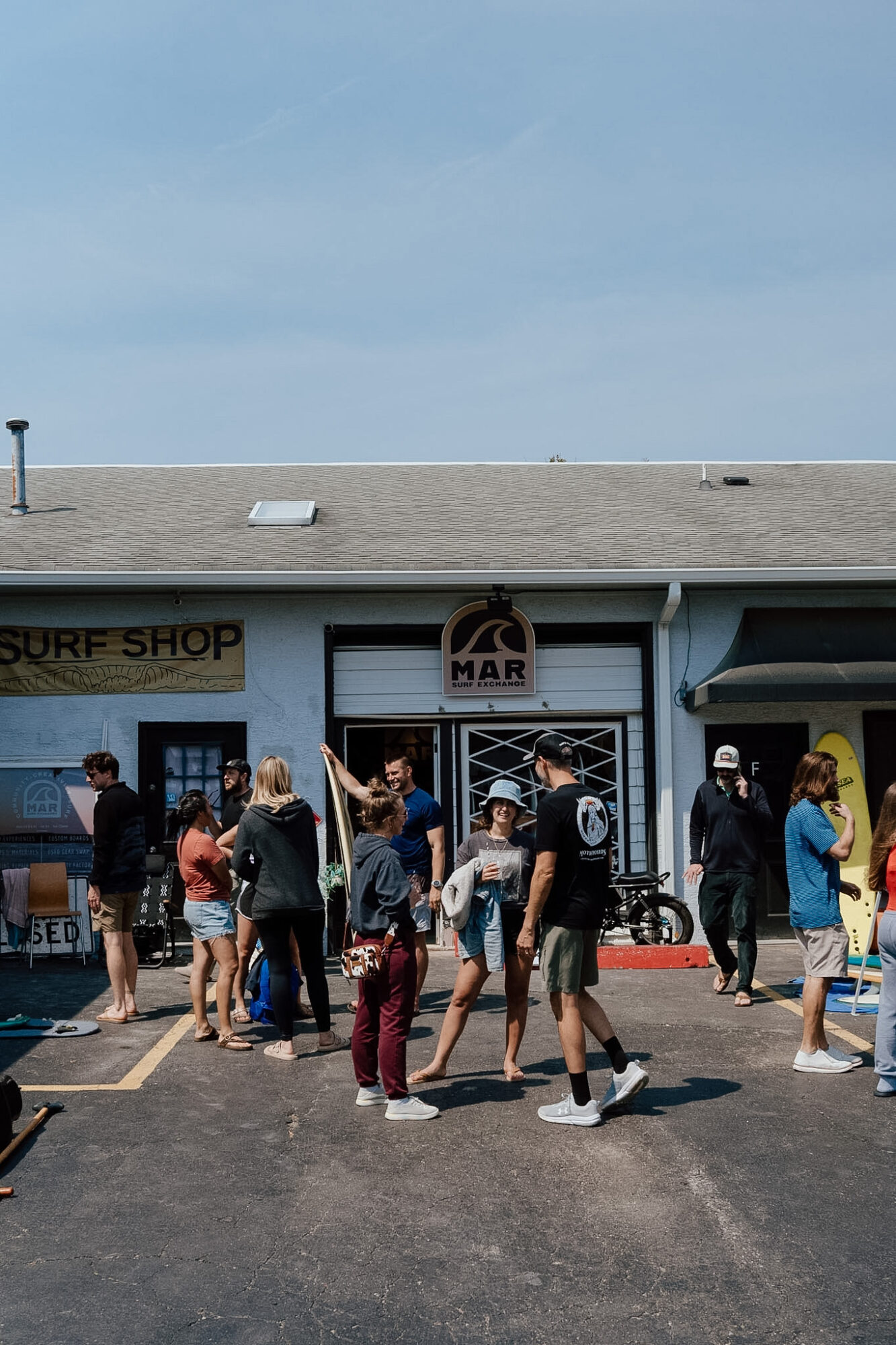 Group of people standing outside a surf shop under a cloudy sky, some talking and walking, storefront with signs and bicycles.