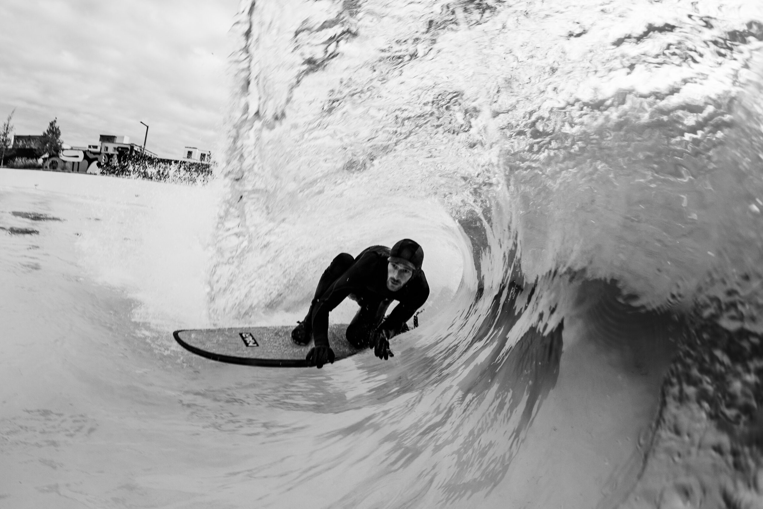 Person surfing inside a wave, crouched on a surfboard, with buildings visible in the background, black and white image.
