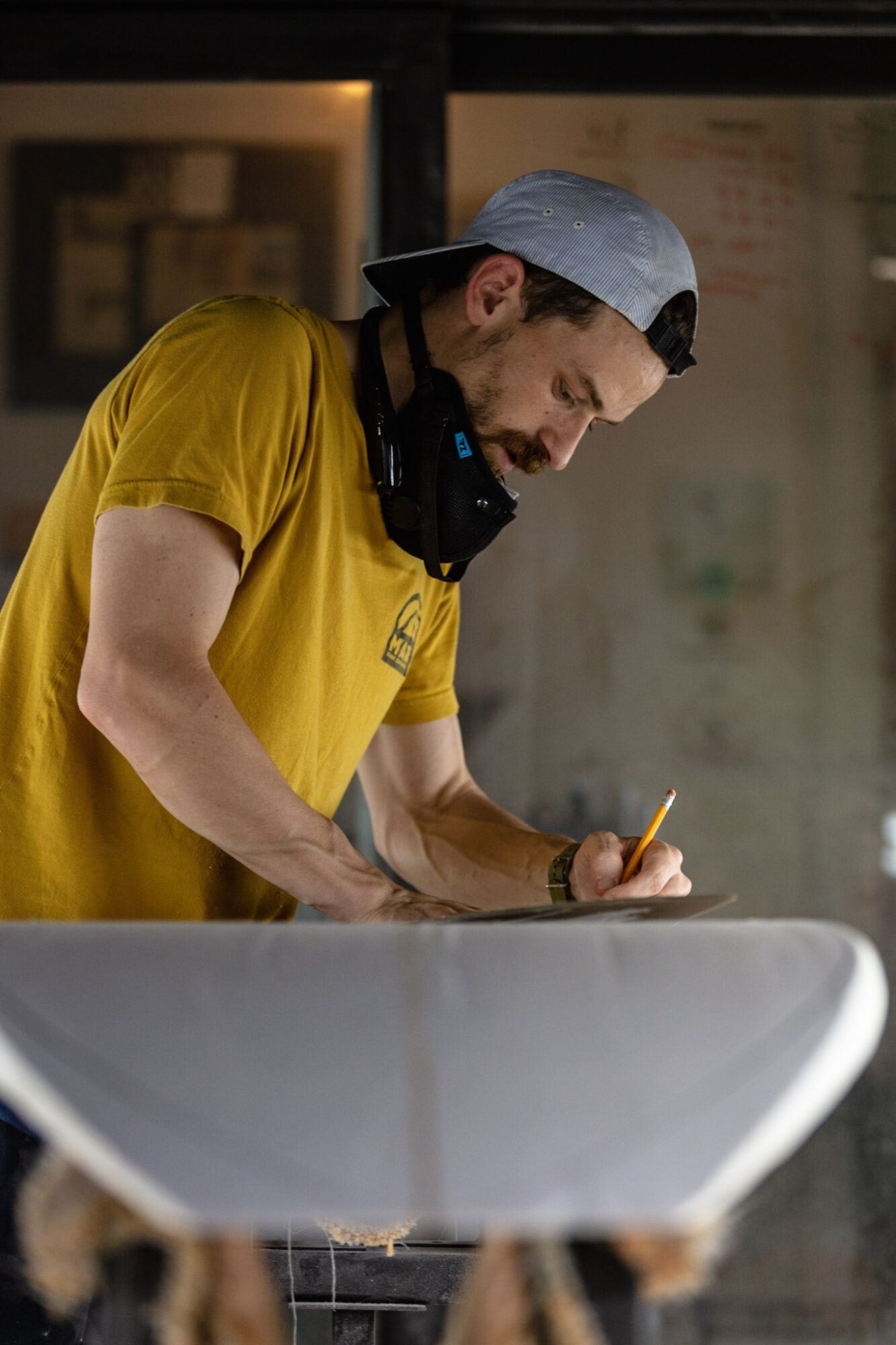 Young man in yellow shirt and gray cap writing on a surface, with headphones around his neck, in an indoor setting.