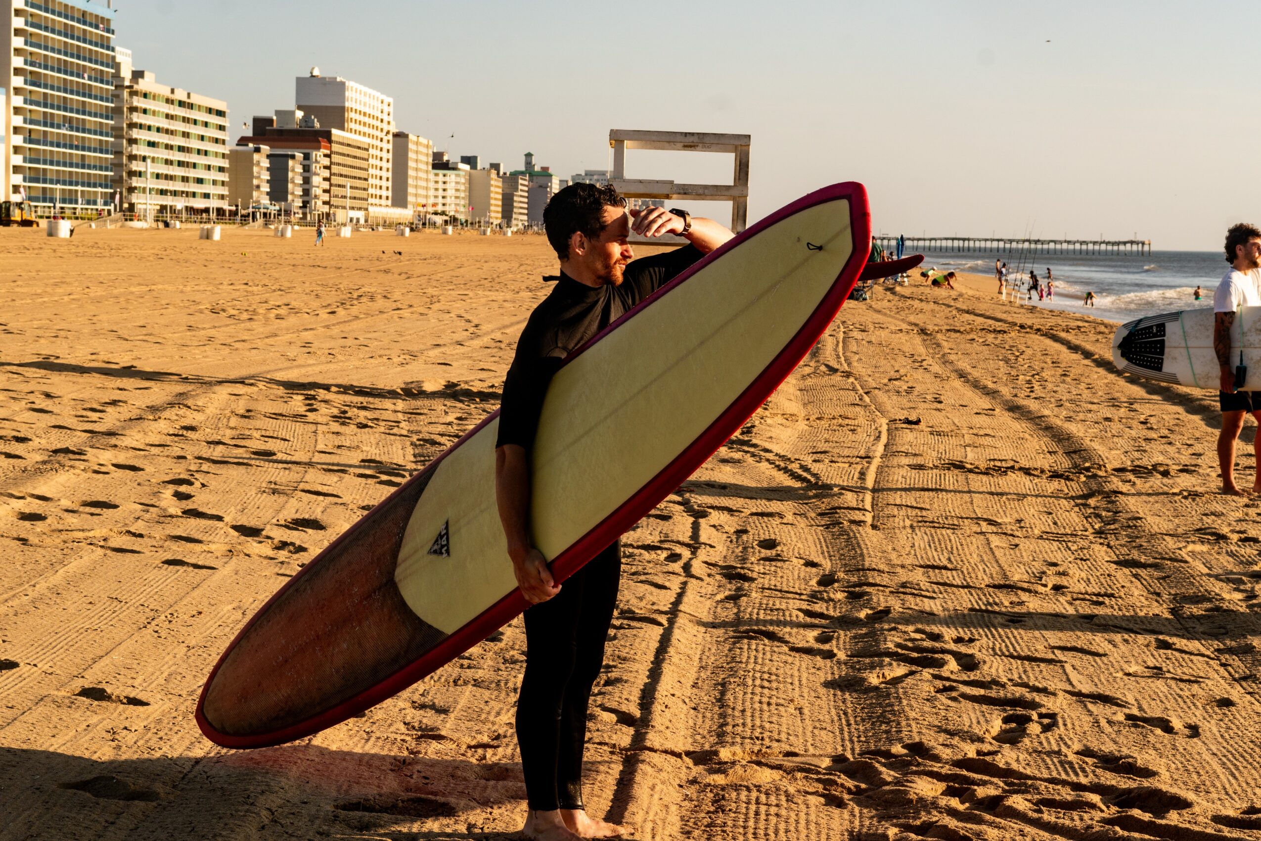 Person holding a surfboard on sandy beach with buildings and ocean in background.