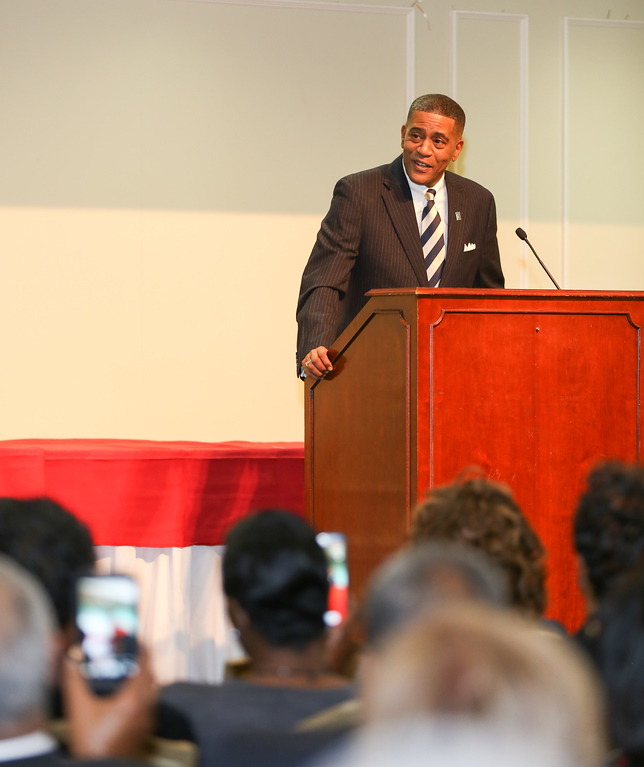 Man in suit speaking at podium during event, audience members taking photos, red tablecloth in background.