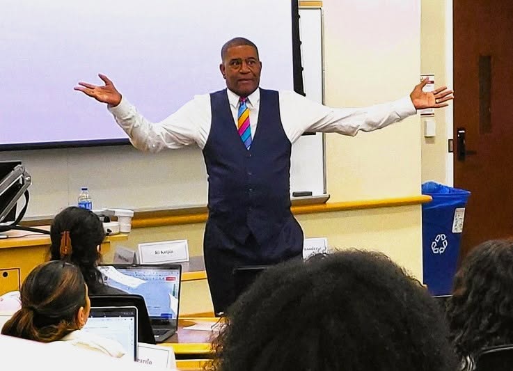 Person standing in front of a classroom with arms outstretched, students using laptops, classroom setting.