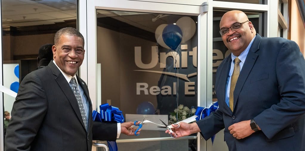 Two men in suits holding a ribbon in front of a building with a United Real Estate sign.