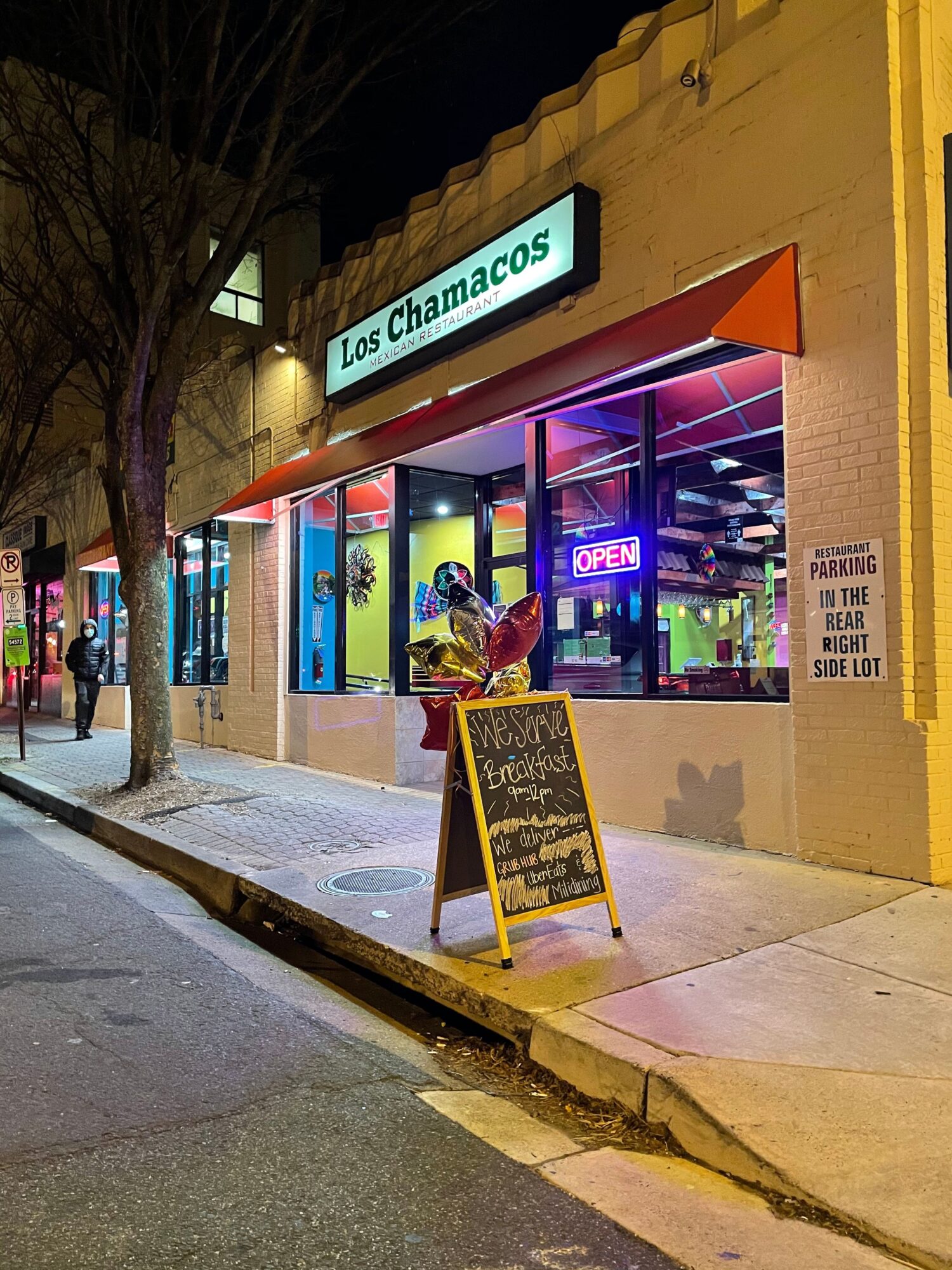 Person standing outside a store with a neon sign, sidewalk, and a chalkboard menu, illuminated by streetlights.