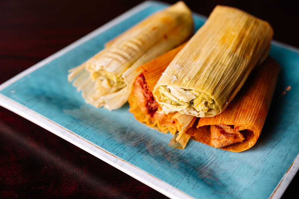 Three tamales on a blue rectangular plate, with one partially unwrapped showing filling, on a dark surface.