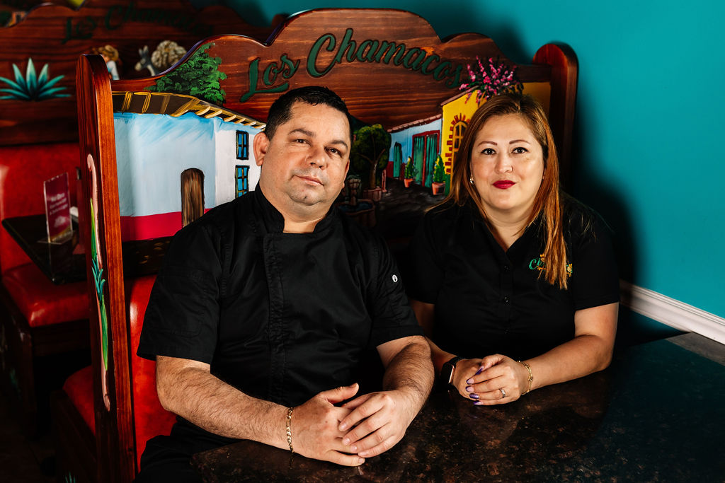 Two people sit at a table in a restaurant with colorful decorations and a sign that reads 