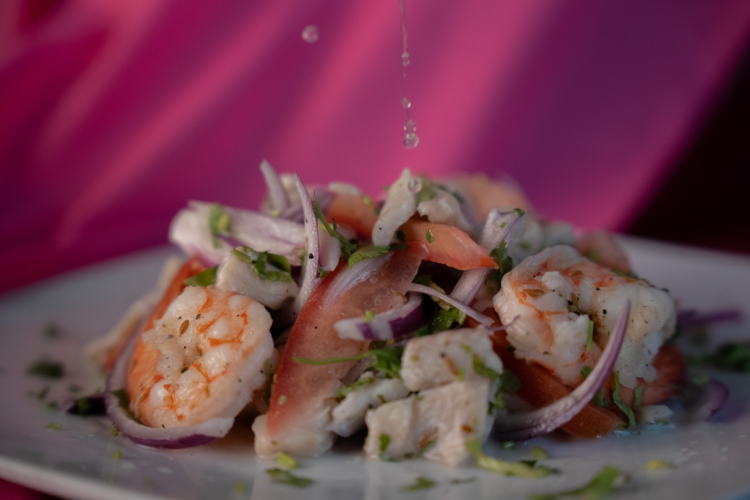 Salad with shrimp, vegetables, and dressing on a white plate, pink background, droplets falling, close-up view.