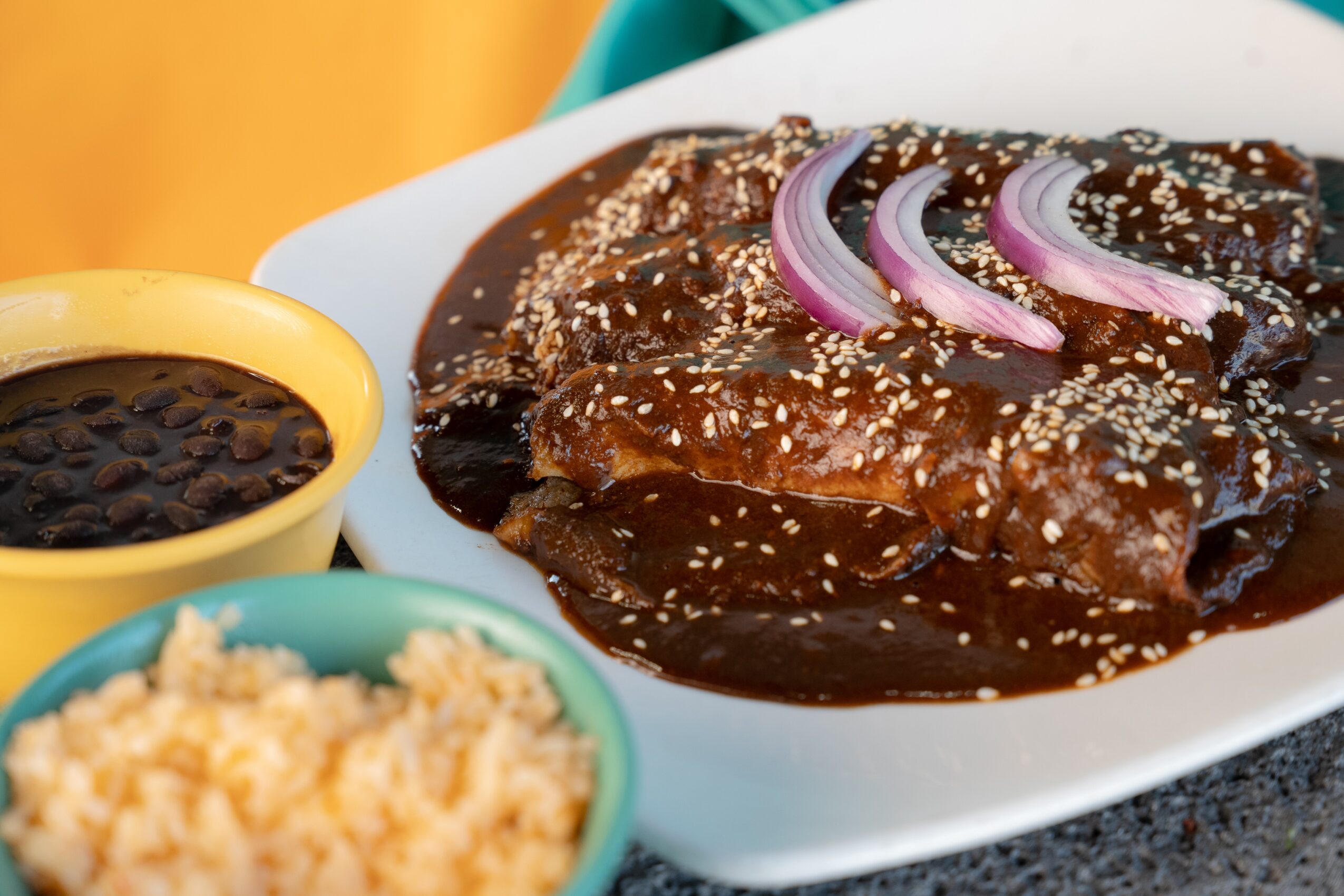 Plate of cooked meat with onion slices and sauce, bowl of rice, bowl of sauce, on a dark surface.