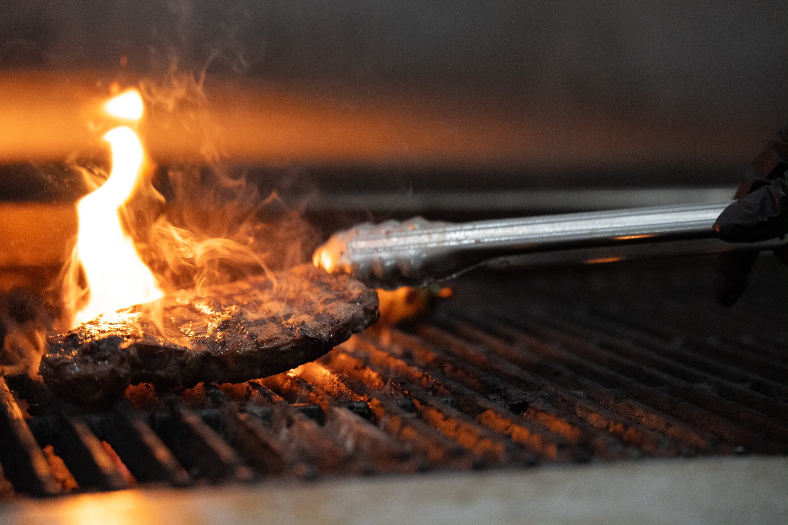 Grill with a piece of meat being cooked over an open flame, tongs holding the meat, grill grates visible.