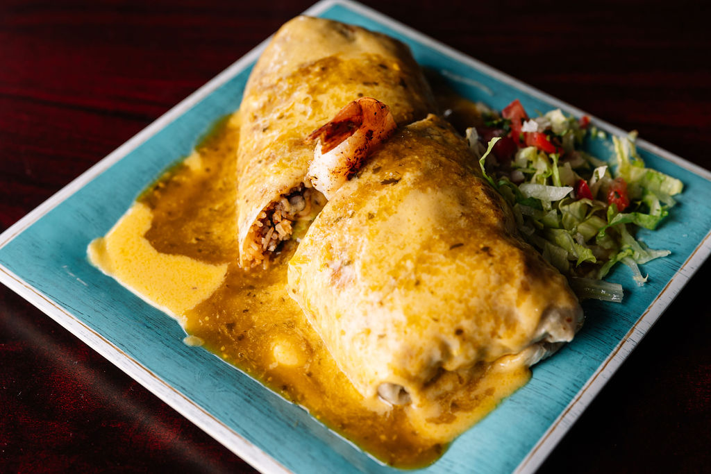 Two burritos covered in sauce on a blue plate with salad, on a dark wooden table.