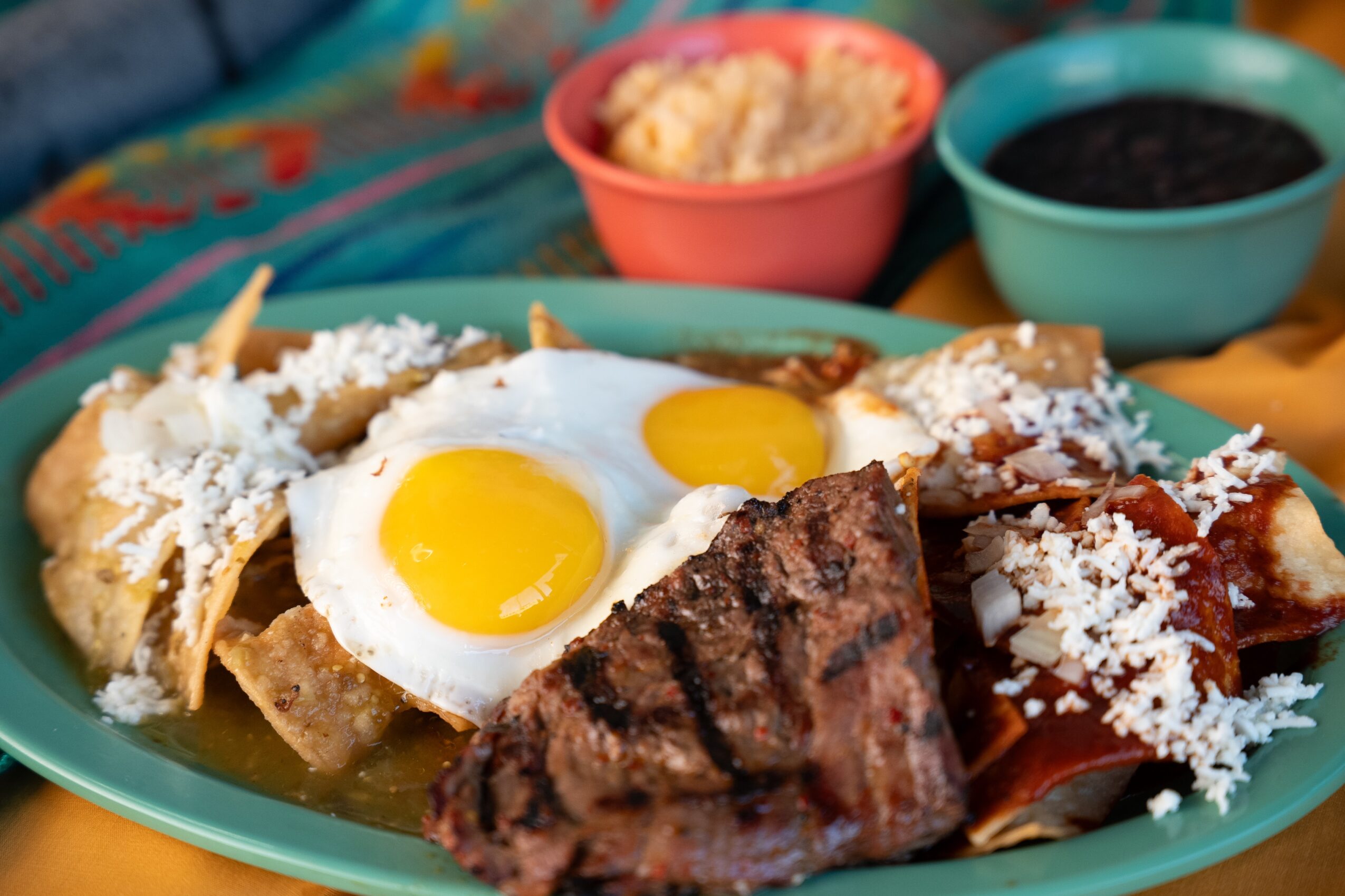 Plate with two fried eggs, grilled meat, cheese, and tortillas, with bowls of salsa and black beans in background.