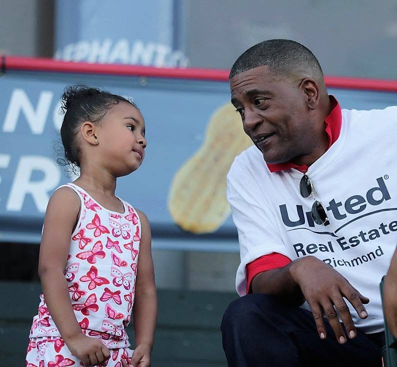 A young girl and a man are engaged in conversation outdoors, with a blurred background featuring a sign and a large ice cream cone image.