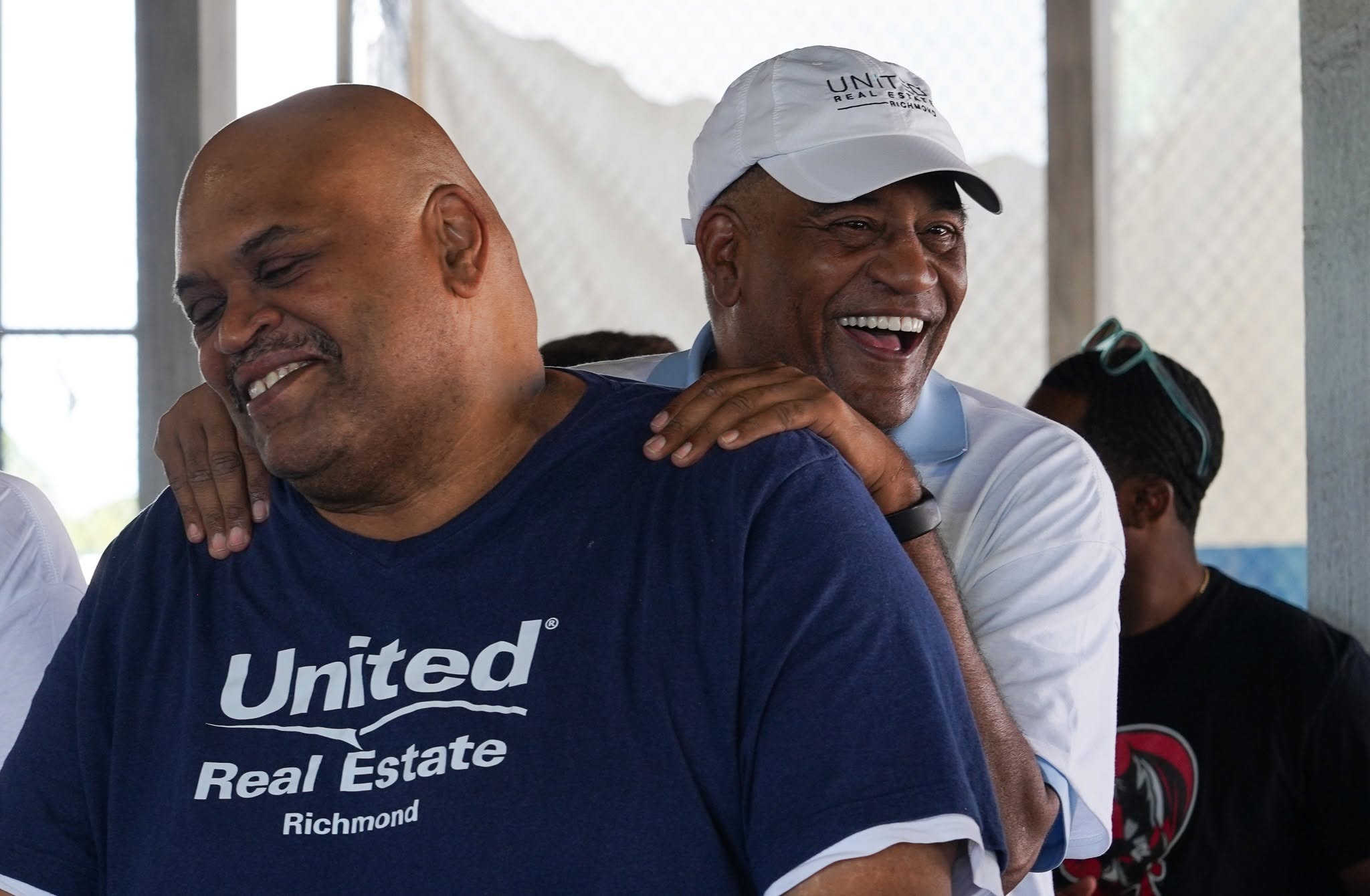 Two men smiling, one with a white cap and the other with a blue shirt, in a bright indoor setting.