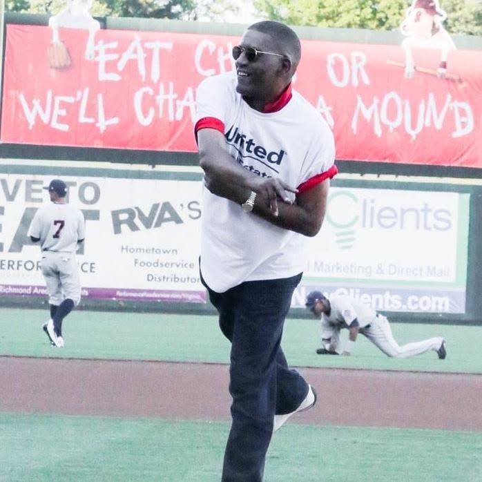 Person smiling, wearing sunglasses and a white sports jersey, standing on a sports field with a banner in the background.