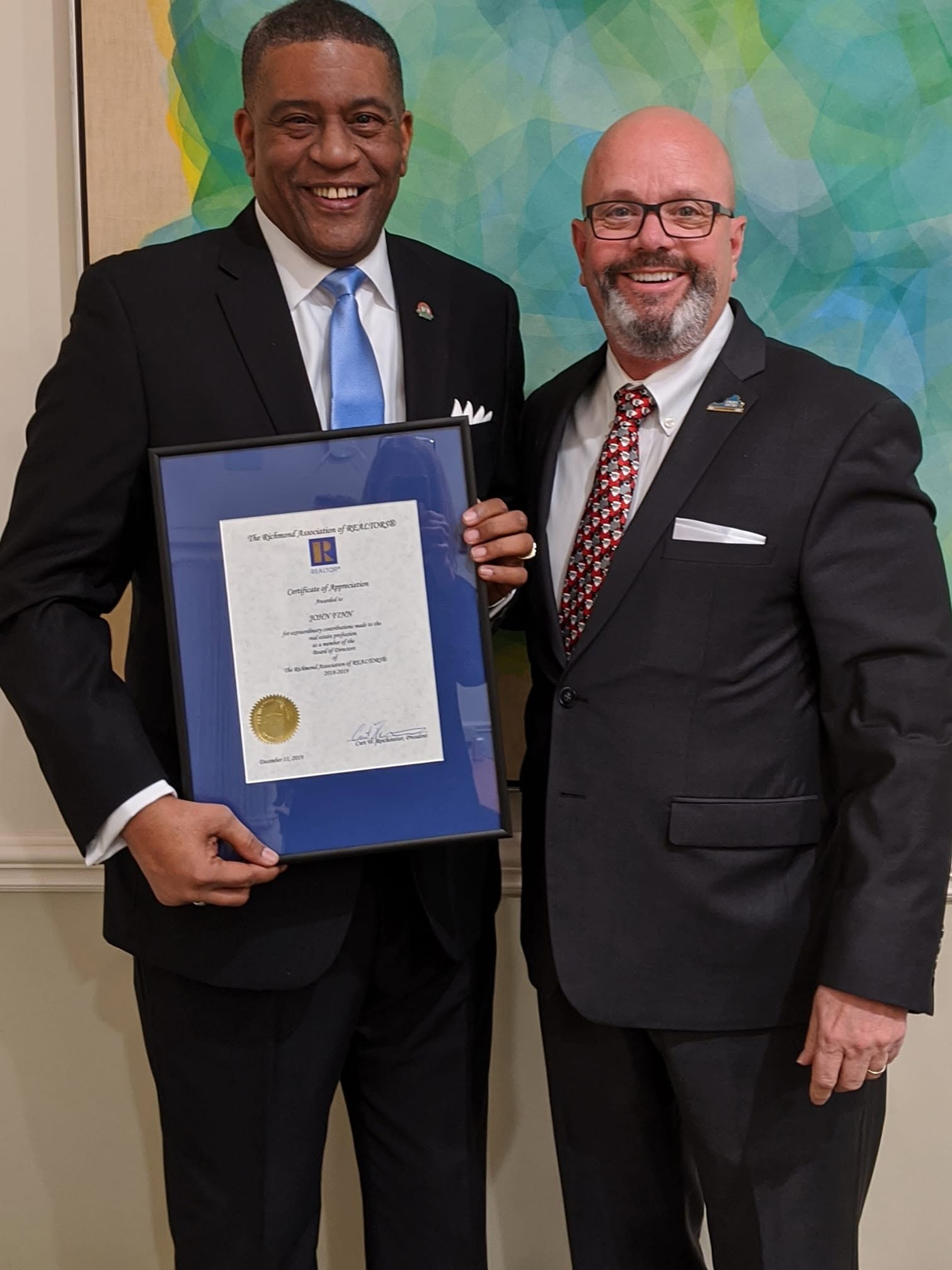 Two men in suits smiling, one holding a framed certificate, standing in front of a colorful background.