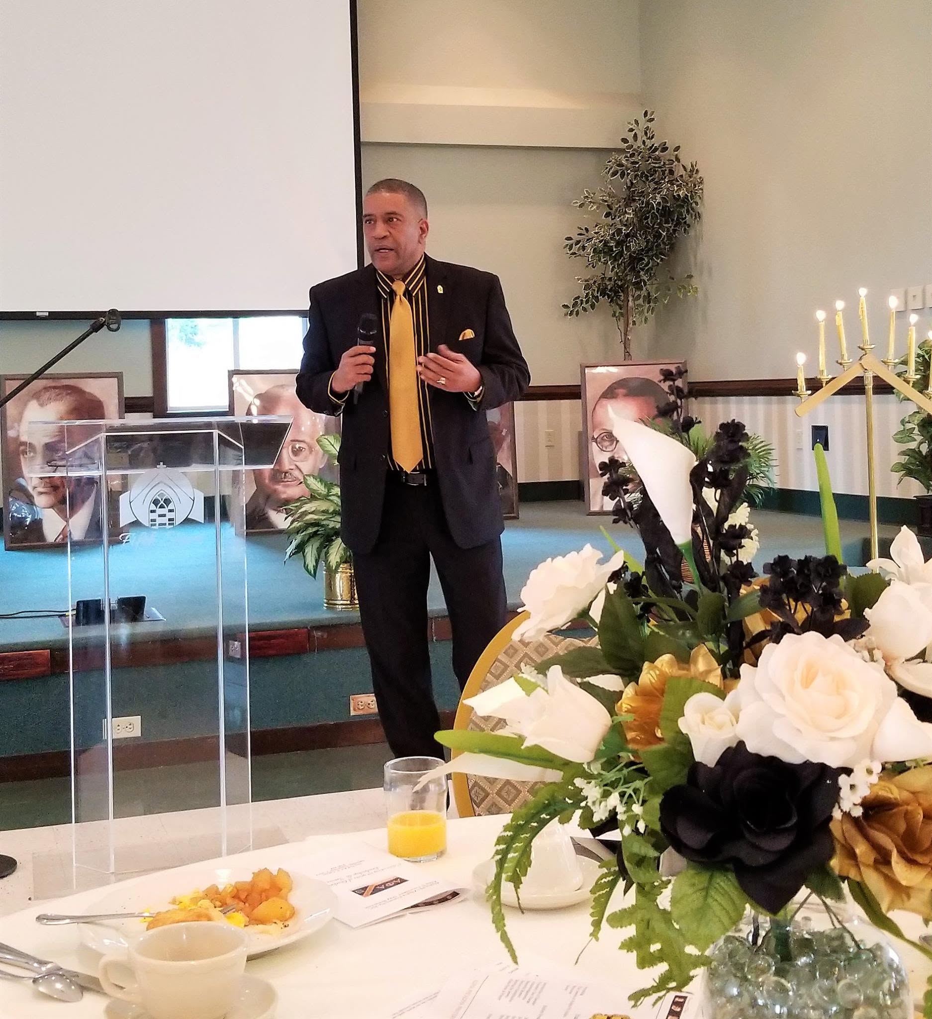 Man in suit speaking at a formal event with floral arrangements and a candelabrum, standing near a transparent lectern.