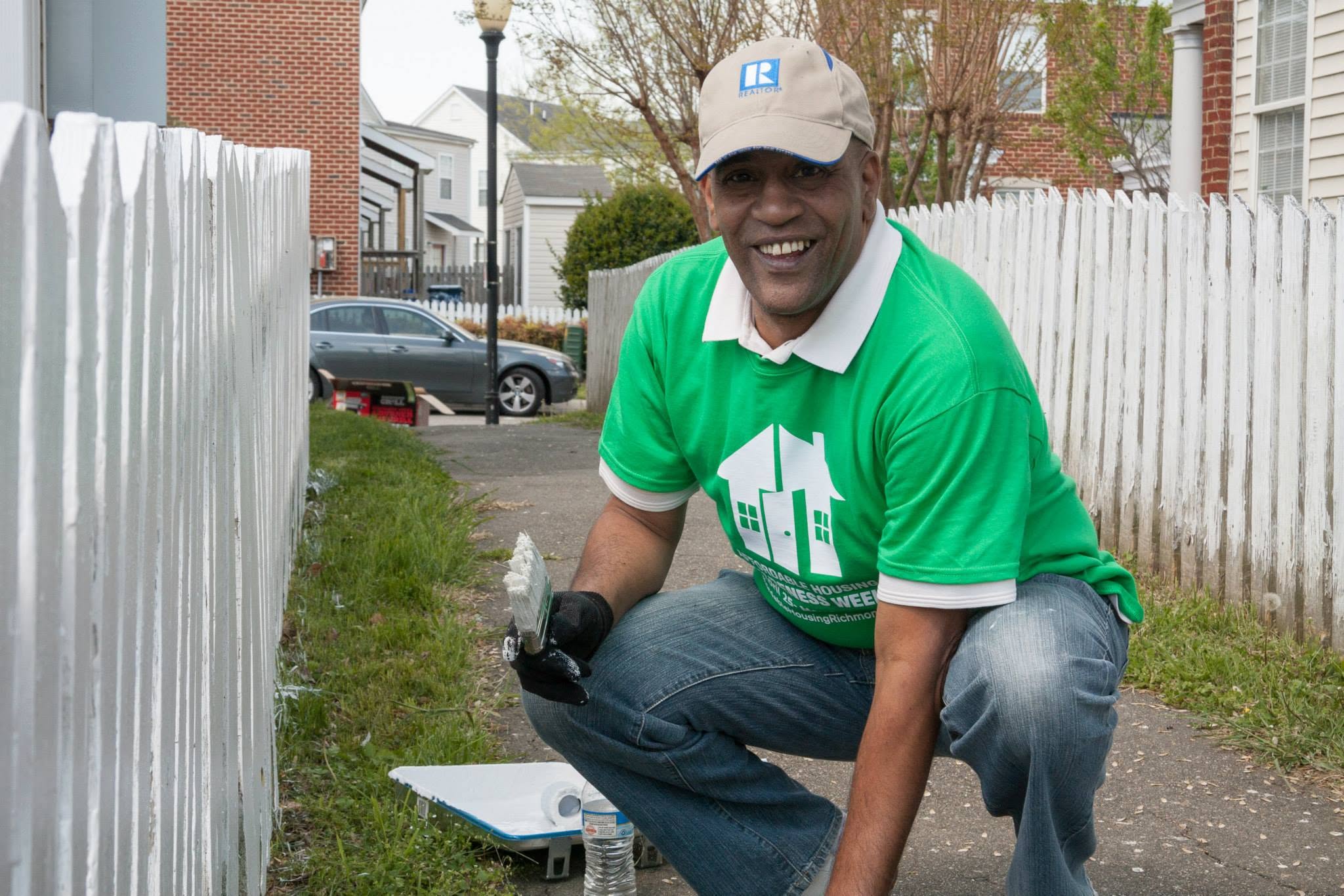 Man crouching outdoors, smiling, wearing a green T-shirt, white cap, and gloves, near a white fence.
