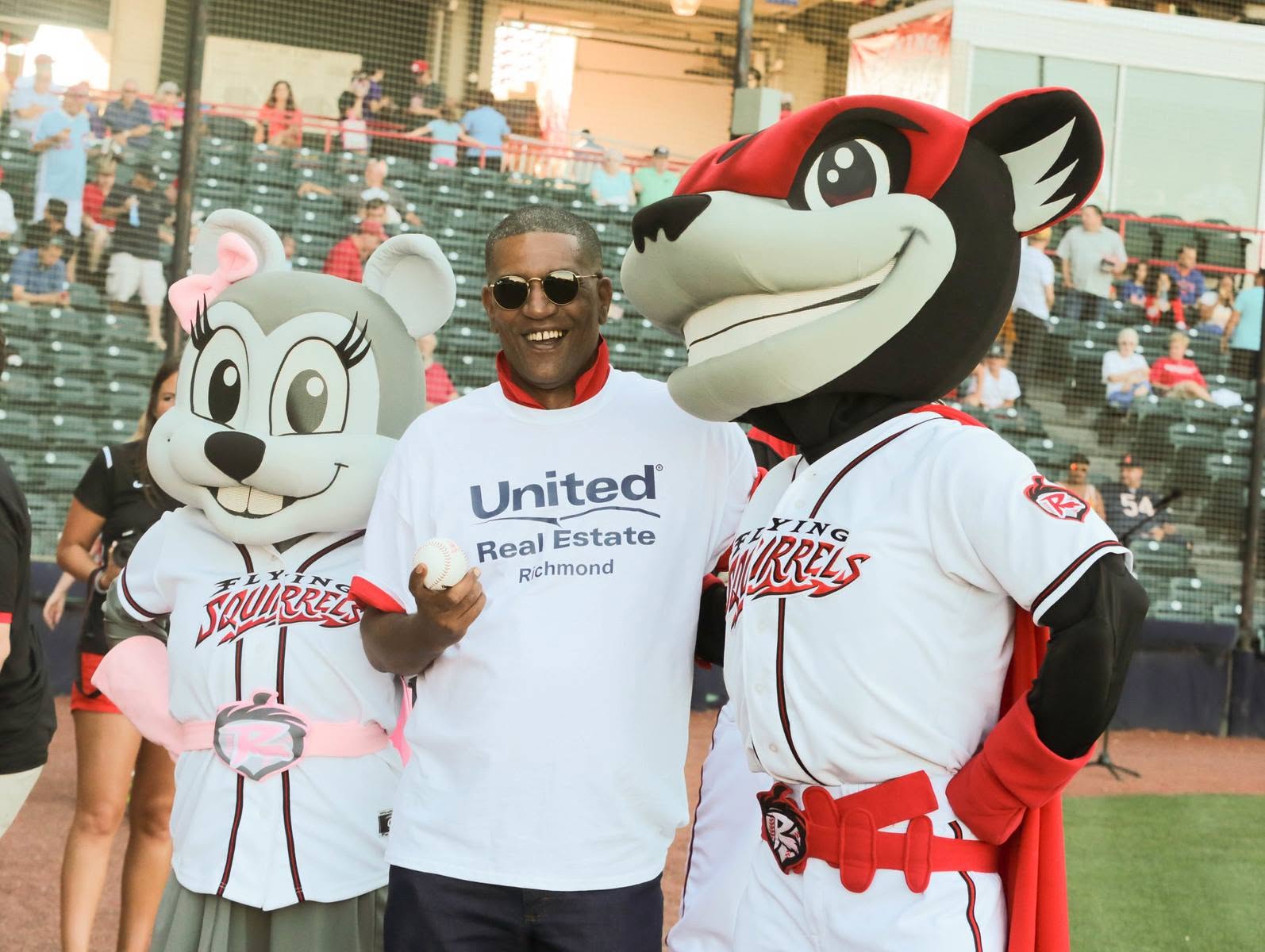 Man in sunglasses standing between two mascots in baseball uniforms at a stadium, smiling and giving a thumbs-up.