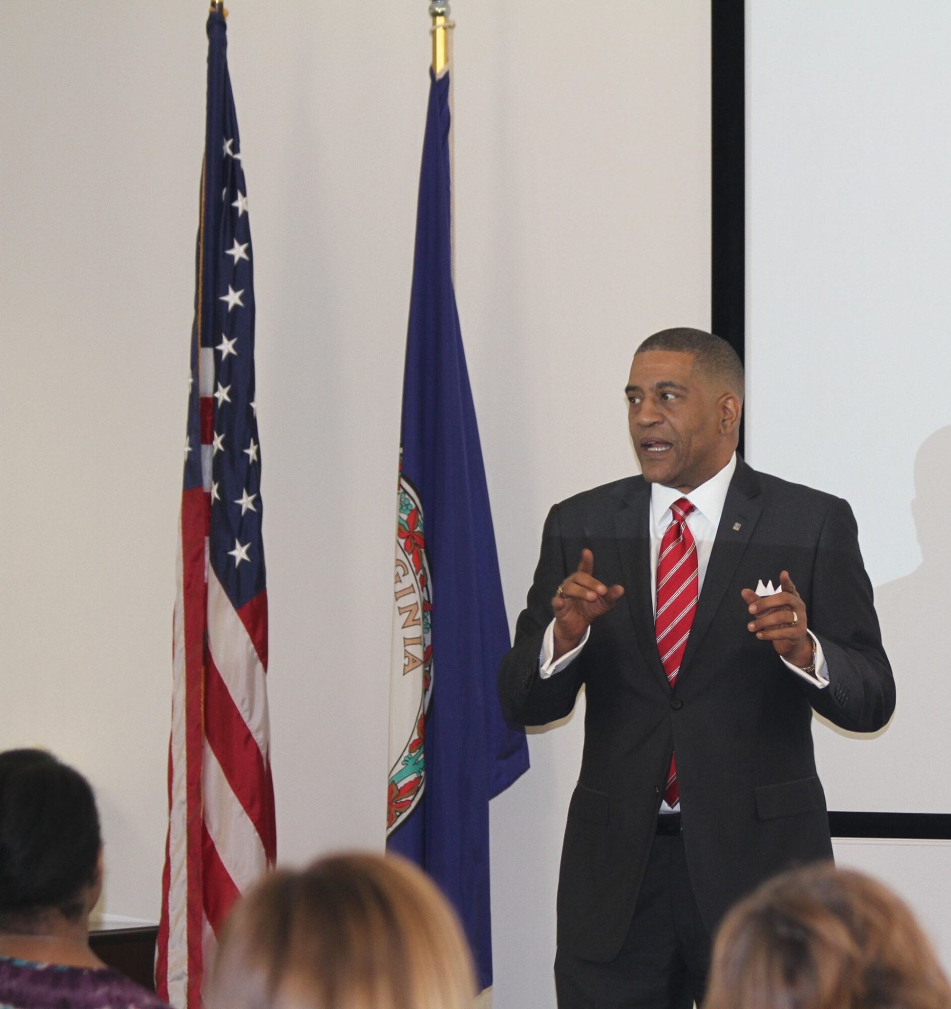 Man in suit speaking to an audience, with American and Virginia flags behind him, in a presentation setting.