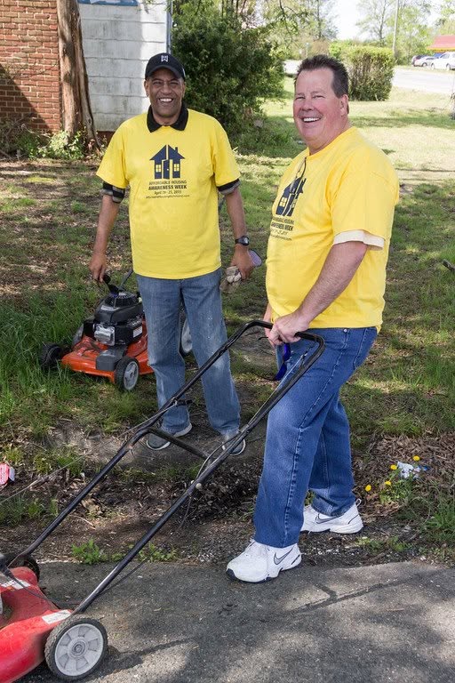 Two men in yellow shirts mowing grass outdoors, smiling, with trees and a building in background.