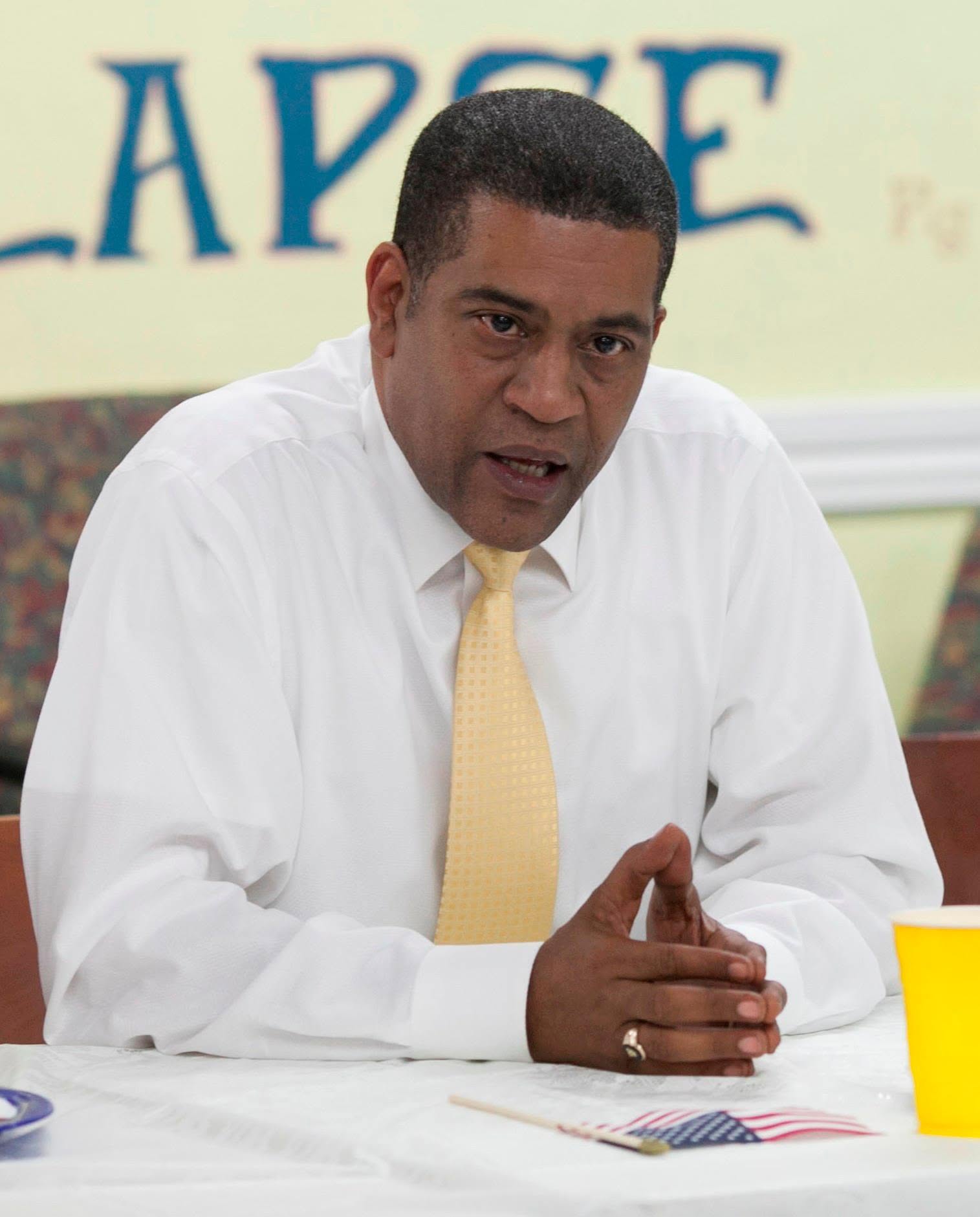 Man in white shirt and yellow tie speaking at a table with a yellow cup, in a room with a sign behind him