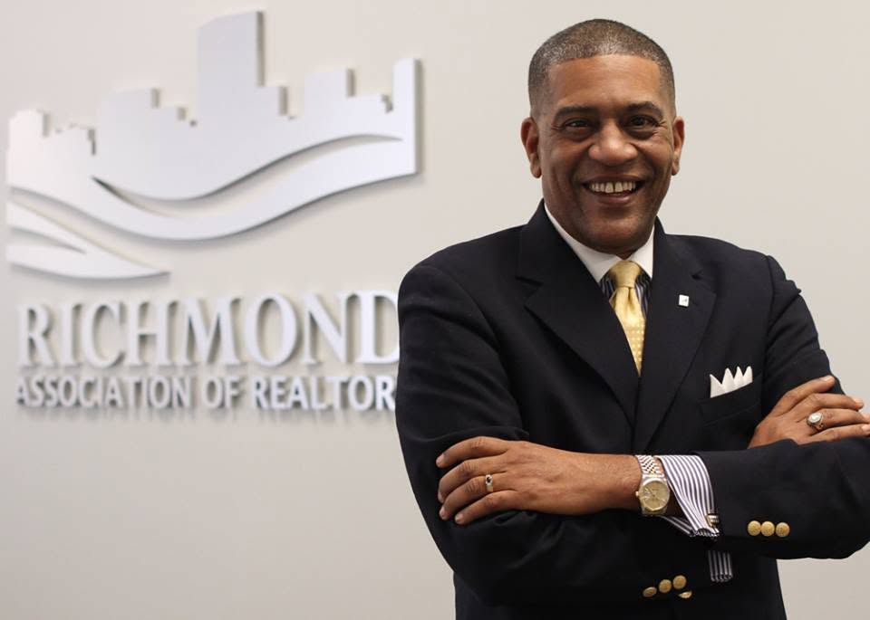 Man in a suit smiling with arms crossed, standing in front of a Richmond Association of Realtors sign.