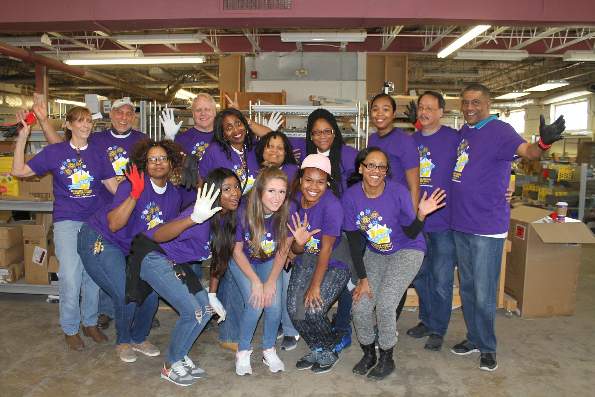 Group of 14 people wearing matching purple shirts, smiling and waving inside a warehouse or factory setting.